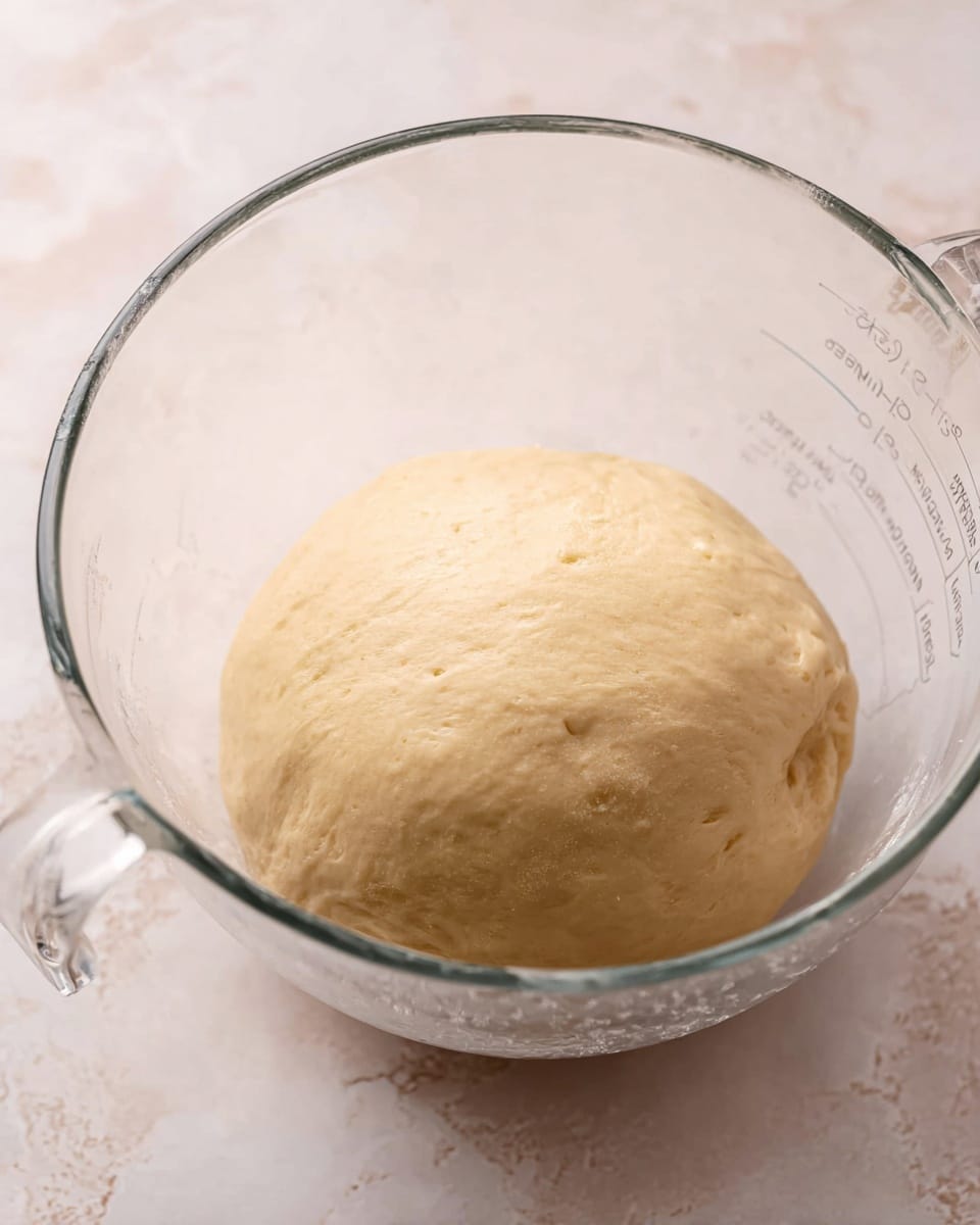 The image shows a smooth, round ball of dough with a light beige color resting at the bottom of a clear glass measuring cup. The dough has a soft and slightly shiny texture with small air bubbles visible on its surface. The glass container is transparent with measurement markings on the side, showing the dough's size compared to the cup. The entire scene is set against a white marbled texture, giving a clean and bright look to the background. photo taken with an iphone --ar 4:5 --v 7