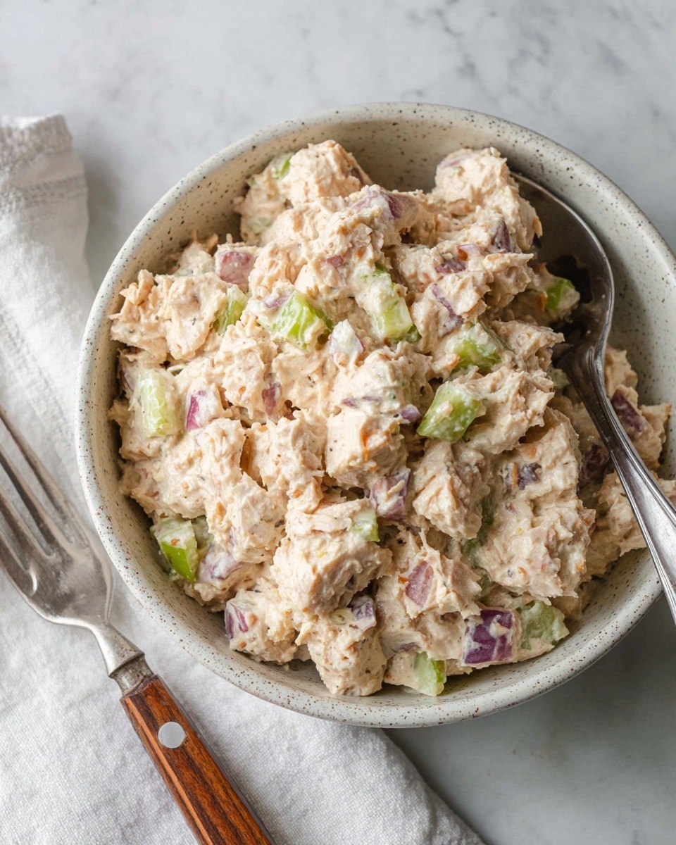 The image shows a close-up of a white speckled bowl filled with a creamy tuna salad mixture. The salad has chunks of light beige tuna combined with small pieces of green celery and purple-red onion, all coated in a light creamy dressing, giving it a slightly lumpy texture. The bowl sits on a white marbled surface with a silver fork with a wooden handle placed partially out of frame on a white cloth beside it. Photo taken with an iphone --ar 4:5 --v 7