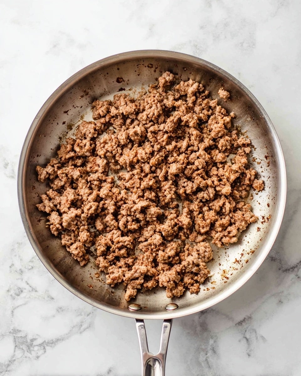 A round silver frying pan filled with cooked ground meat spread unevenly inside, with some parts browned more than others, resting on a white marbled surface. The meat pieces are small, crumbly, and vary in color from light to dark brown. The pan handle is visible and shiny. Photo taken with an iphone --ar 4:5 --v 7