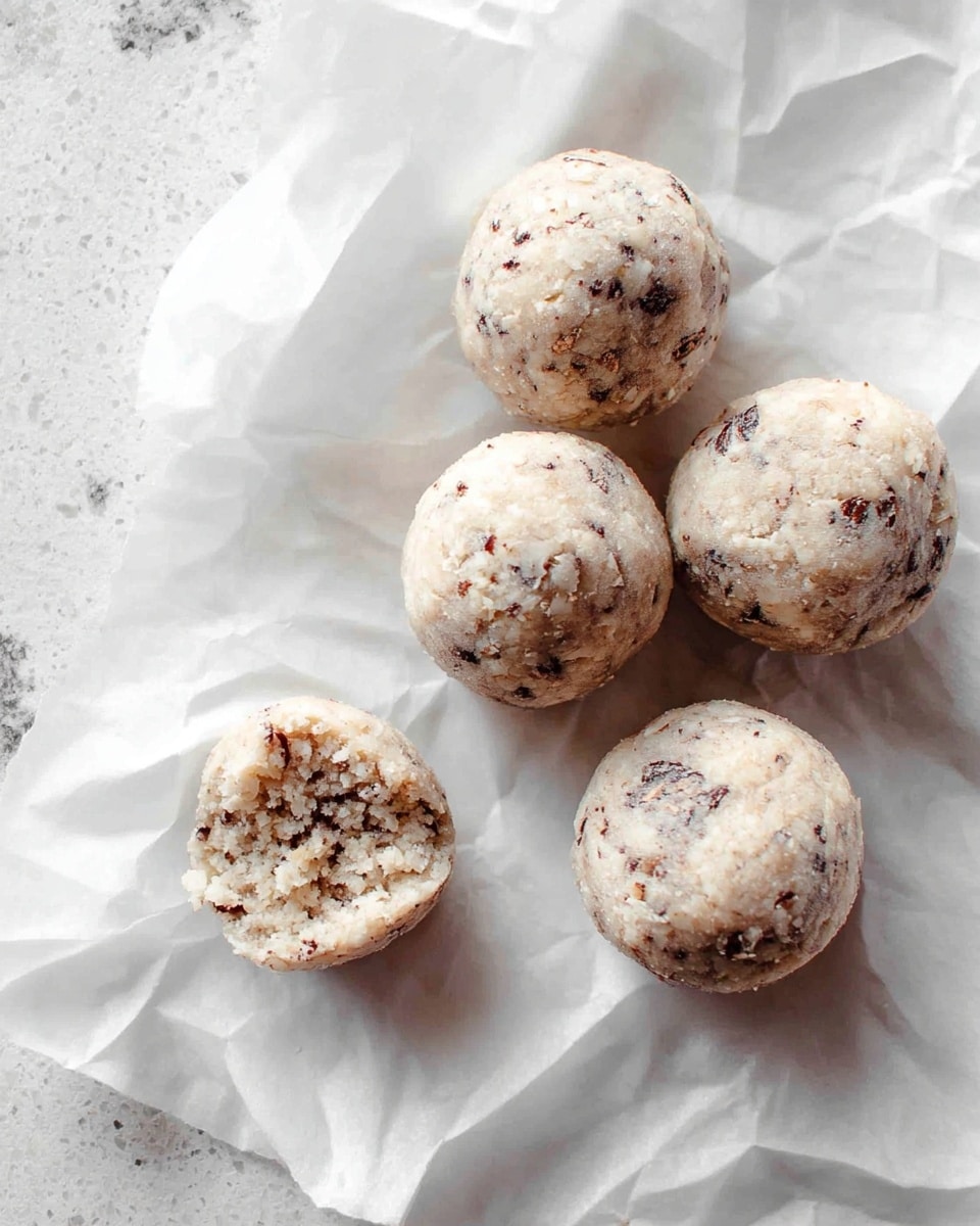 Five small round balls of light beige dough with small dark specks are placed on crumpled white parchment paper. One of the balls is bitten, showing a crumbly and textured inside mixed with the same dark flecks. The background features a white marbled surface, giving a clean and soft look. photo taken with an iphone --ar 4:5 --v 7