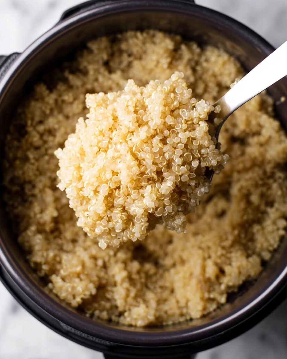 A close-up view of a black electric pot filled with cooked quinoa, showing a white plastic spoon lifting a portion of the fluffy, light tan quinoa grains. The quinoa has a soft, slightly grainy texture with small, curled tails visible on many grains. The background surface is a white marbled texture. Photo taken with an iphone --ar 4:5 --v 7