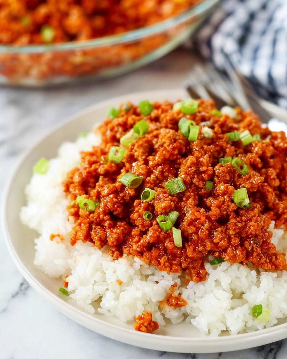 A close-up view of a white plate filled with a base layer of fluffy white rice. On top of the rice is a thick layer of cooked ground meat in a rich red-orange sauce, with small green onion pieces scattered evenly across the meat, adding a touch of green. The dish appears moist with a slightly shiny texture, showing bits of sauce clinging to the meat and rice. The plate is set on a white marbled surface with a blurred glass bowl of the same dish in the background. photo taken with an iphone --ar 4:5 --v 7