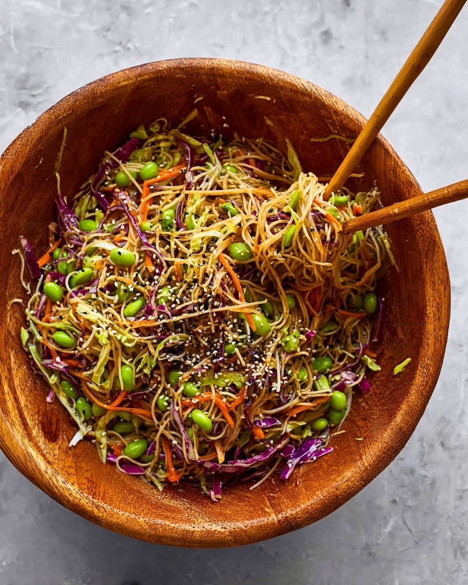 A large wooden bowl filled with a colorful noodle salad sits on a white marbled surface. The dish has a base of thin, light brown noodles mixed with bright green edamame beans scattered throughout. There are finely shredded vegetables layered in, including pale green cabbage, purple cabbage, and thin strips of orange carrot, all mixed evenly with the noodles. The dish is topped with small black and white sesame seeds sprinkled across the surface. Two wooden chopsticks are placed in the bowl, one lifting a small bunch of noodles. photo taken with an iphone --ar 4:5 --v 7