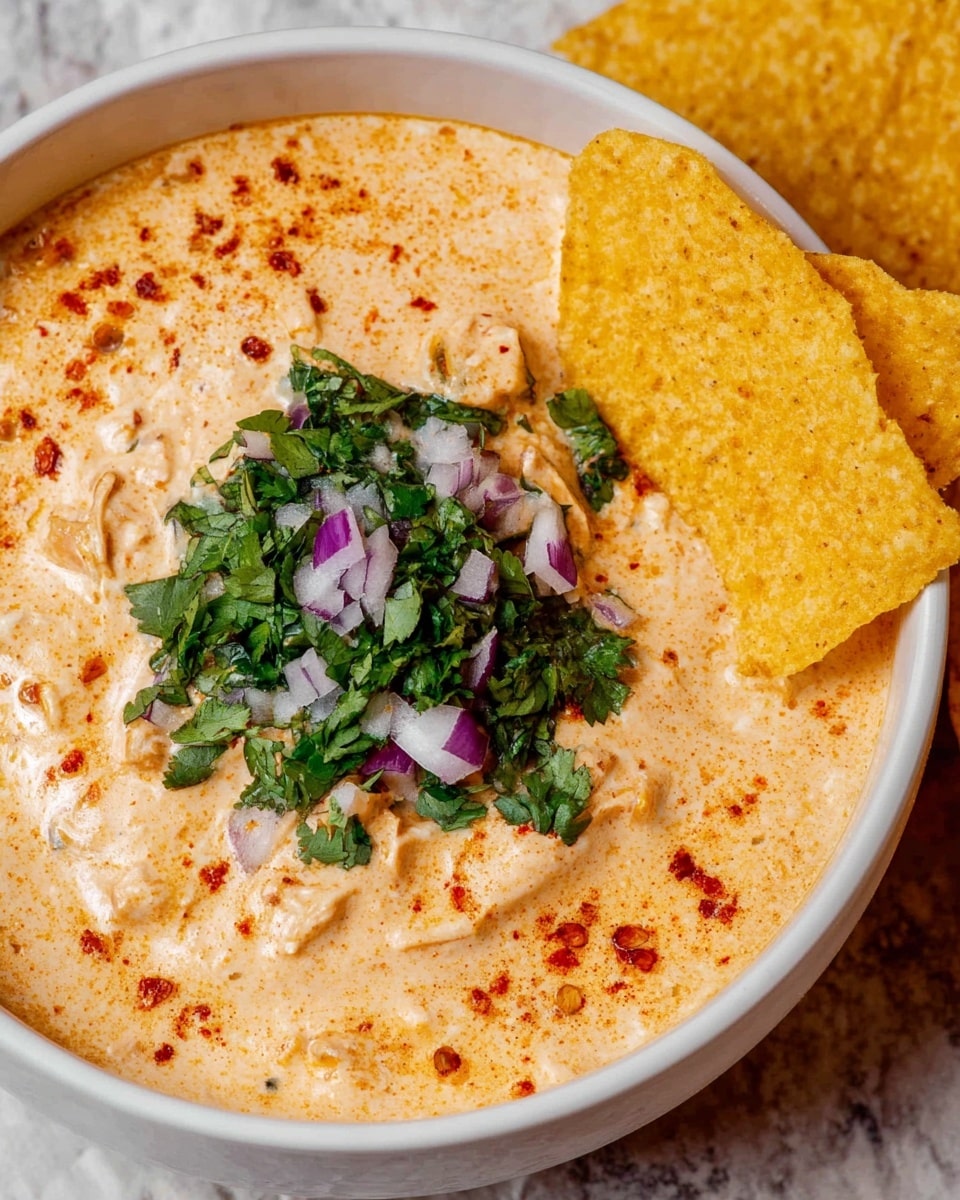 A white bowl filled with a creamy, light orange dip sprinkled with small red specks, topped in the center with chopped fresh green cilantro and diced pieces of purple and white onion. Two yellow tortilla chips with a slightly toasted texture rest inside the bowl on the right side, partially dipped in the creamy mixture. The bowl sits on a surface with a white marbled texture. photo taken with an iphone --ar 4:5 --v 7