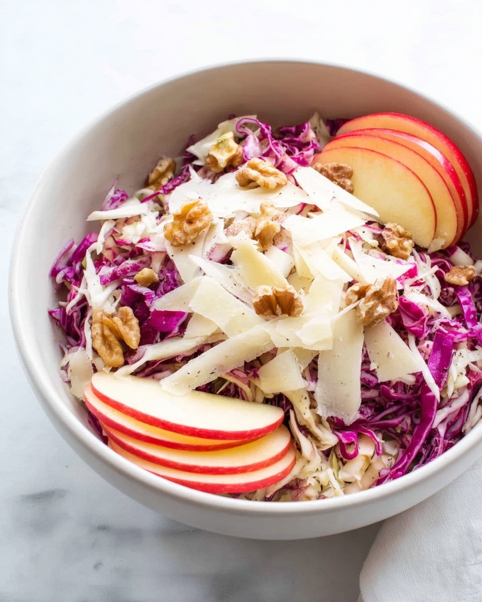 The image shows a white bowl filled with a colorful layered salad placed on a white marbled surface. The bottom layer is shredded white and purple cabbage, giving a mix of light and deep purple colors with a crisp texture. On top of the cabbage, there are thin slices of red apples arranged around the edge, adding a bright red color with a smooth texture. Scattered over the apples and cabbage are small pieces of light brown walnuts, adding a rough texture and crunchy look. Thin white shavings, likely cheese or a similar ingredient, are spread across the top with a slightly translucent, smooth texture. The salad looks fresh, colorful, and ready to eat. Photo taken with an iphone --ar 4:5 --v 7