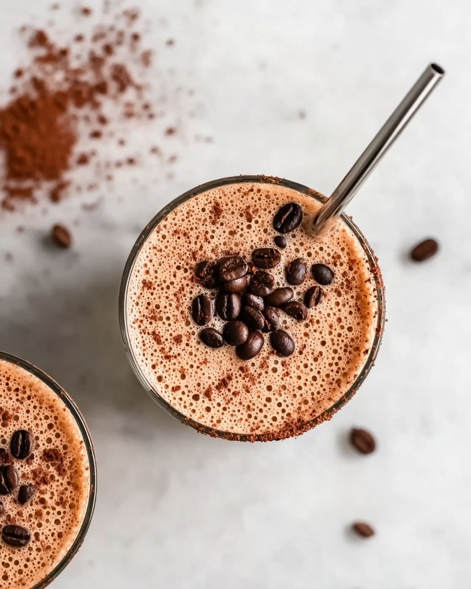 The image shows a top view of two clear glasses filled with a light brown frothy drink. The drink has a thick layer of small bubbles on top, sprinkled with dark brown cocoa powder and whole dark coffee beans scattered unevenly. A metal straw is placed in the right glass, extending upward and slightly to the right. The glasses sit on a white marbled surface with some cocoa powder and coffee beans scattered around them, and a woman's hand gently holds the left glass. Photo taken with an iphone --ar 4:5 --v 7