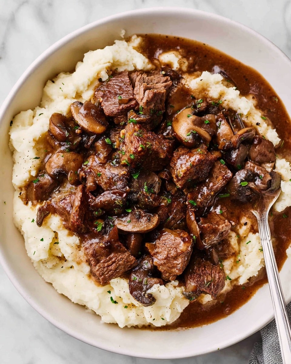 A white bowl is filled with two main layers; the bottom layer is creamy mashed potatoes, soft and white with a smooth texture, spread evenly inside the bowl. The top layer shows rich brown pieces of beef and dark mushrooms cooked together in a thick brown gravy, scattered generously over the mashed potatoes. The beef chunks are tender and textured, and the mushrooms add a darker, slightly glossy contrast. Small green herb bits are sprinkled on top for color. A silver fork is partially placed into the dish on the right side. The bowl is set on a white marbled surface. photo taken with an iphone --ar 4:5 --v 7