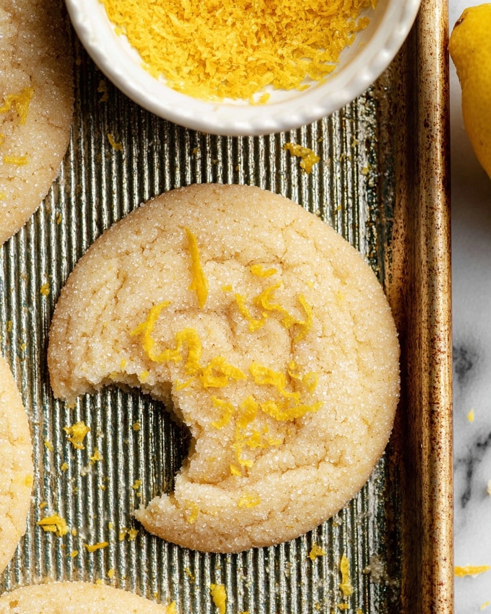 A close-up view of a round, light beige cookie with a rough, sugary texture, sprinkled with small bright yellow lemon zest pieces on top. The cookie has a bite taken from its upper left side, revealing a soft inside. It sits on a metallic baking tray with a striped pattern. To the upper left, there is a white bowl filled with finely grated yellow lemon zest, and on the lower right side, a whole yellow lemon is partially visible. The surface below the tray is a white marbled texture. photo taken with an iphone --ar 4:5 --v 7