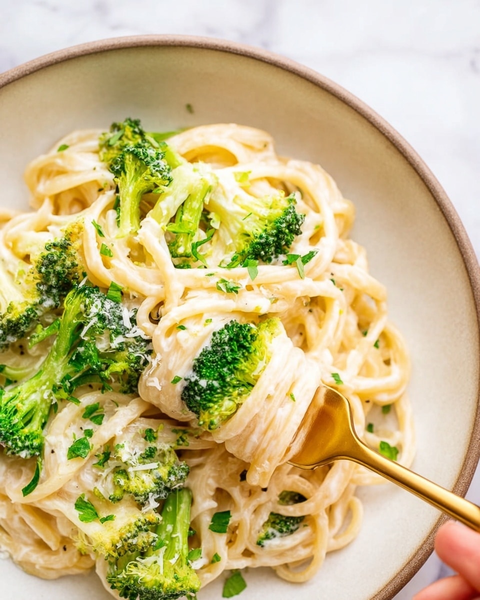 The image shows a close-up of creamy white pasta noodles mixed with bright green broccoli florets in a white bowl. The noodles are thick and coated with a smooth, pale sauce, with small pieces of chopped green herbs sprinkled on top. A shiny golden fork is twirling some noodles in the center, and a woman's hand is holding the fork from the right side. The bowl sits on a white marbled surface, giving the scene a clean and bright look. photo taken with an iphone --ar 4:5 --v 7