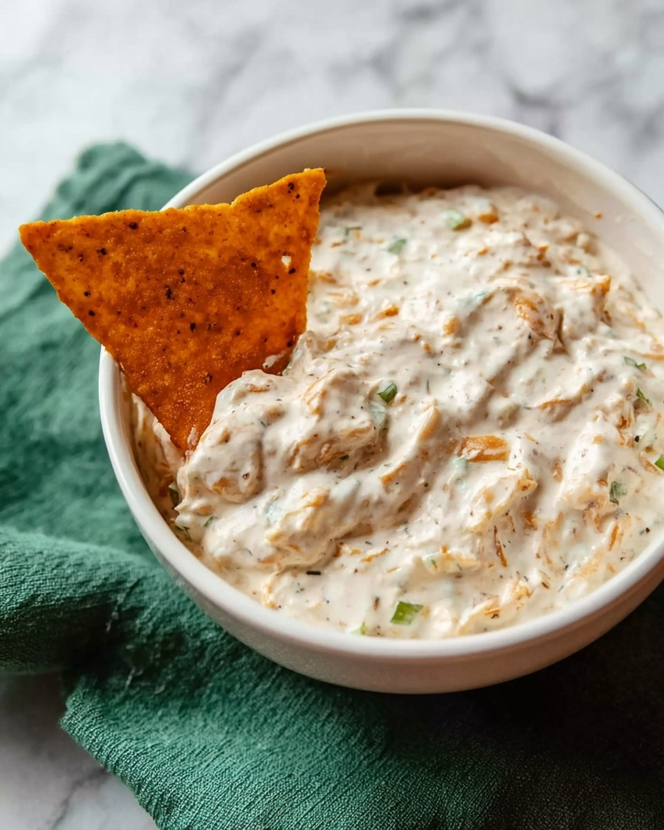 A white bowl filled with creamy white dip that has small green and light brown bits mixed inside, giving it texture. A triangular chip with orange and black specks is dipped into the thick dip, covered halfway. The bowl sits on a white marbled surface, and a green cloth is partially visible underneath the bowl. Photo taken with an iphone --ar 4:5 --v 7