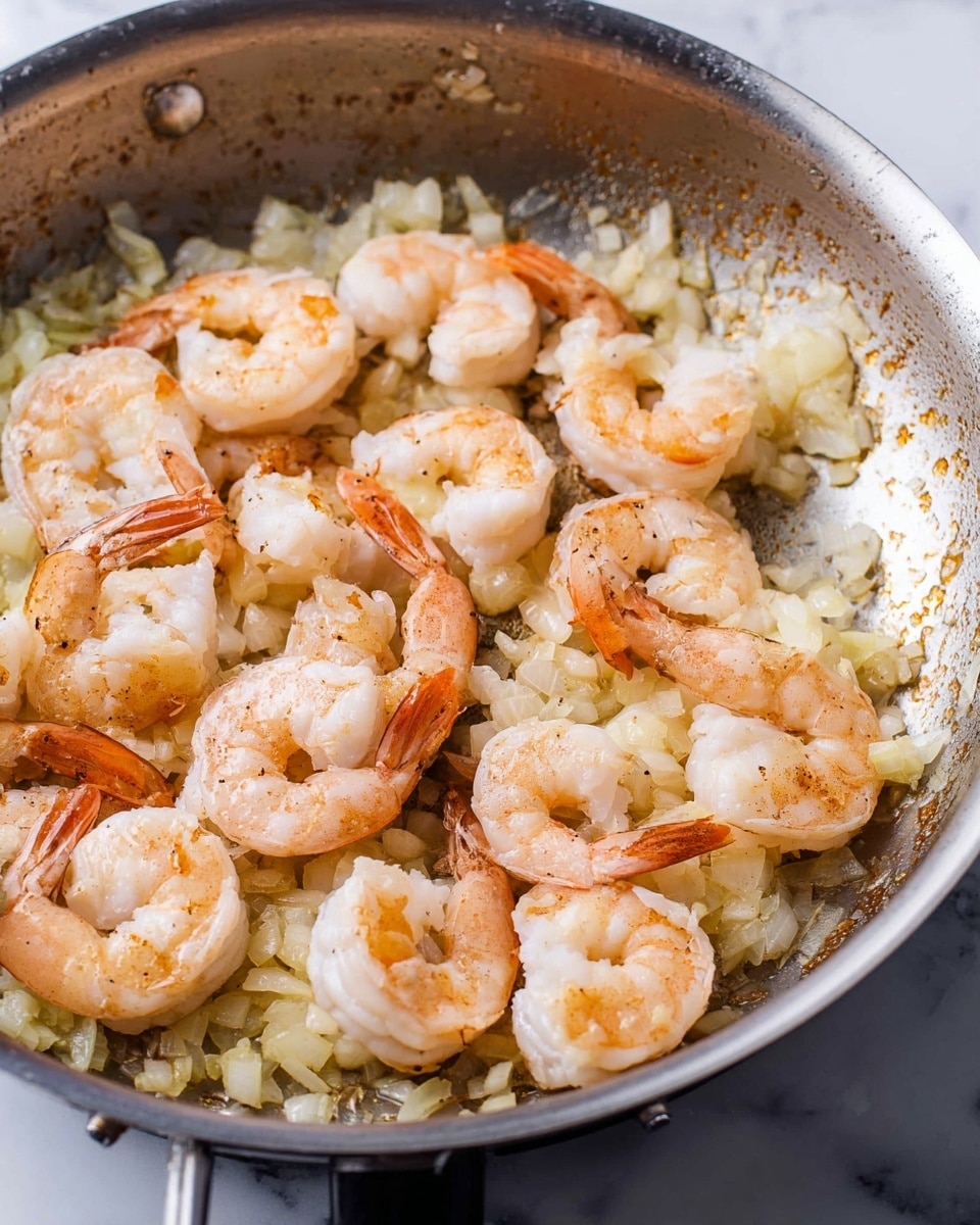 The image shows a close-up of a silver pan on a white marbled surface, filled with shrimp cooking over a layer of small chopped onions. The shrimp are light pink with some parts still white, showing signs of light cooking with slight browning on some edges. The onions underneath are soft and pale yellow, spread evenly to cover the pan bottom. The pan has a shiny metallic texture with some brown spots from cooking. photo taken with an iphone --ar 4:5 --v 7