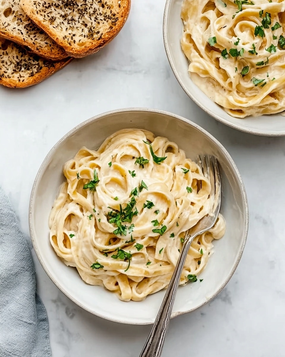 The image shows two white bowls filled with creamy fettuccine pasta. Each bowl has about three layers of thick pasta with a smooth, light beige sauce covering the noodles completely. Small green herb bits are scattered on top of the pasta for contrast. A silver fork sits in the front bowl on the right side, slightly resting on the edge of the bowl. To the upper left, two slices of toasted bread with seeds are placed on the white marbled surface. The whole scene is bright and clean, with natural light enhancing the soft textures of the sauce and pasta. photo taken with an iphone --ar 4:5 --v 7