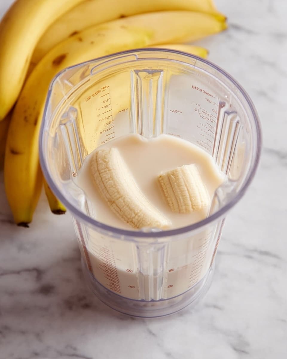 A clear blender container is shown with two peeled banana pieces inside, partly submerged in a light cream-colored liquid, likely milk. The blender is placed on a white marbled surface, with a bunch of yellow bananas resting nearby. The clear blender has measurement markings on its sides and a ribbed texture. The image is bright and clean-looking, focusing on the simple ingredients before blending photo taken with an iphone --ar 4:5 --v 7