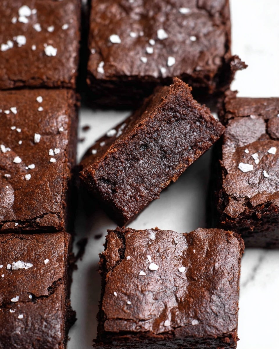 This image shows a batch of dark brown brownies arranged in a grid shape on a white marbled surface. The brownies have a slightly cracked and glossy top with a few white salt crystals sprinkled over them. One brownie piece is lifted slightly above the others, revealing a dense and moist inside texture that looks rich and chocolatey. The edges of the brownies are firm and squared, showing a neat cut between each piece. photo taken with an iphone --ar 4:5 --v 7