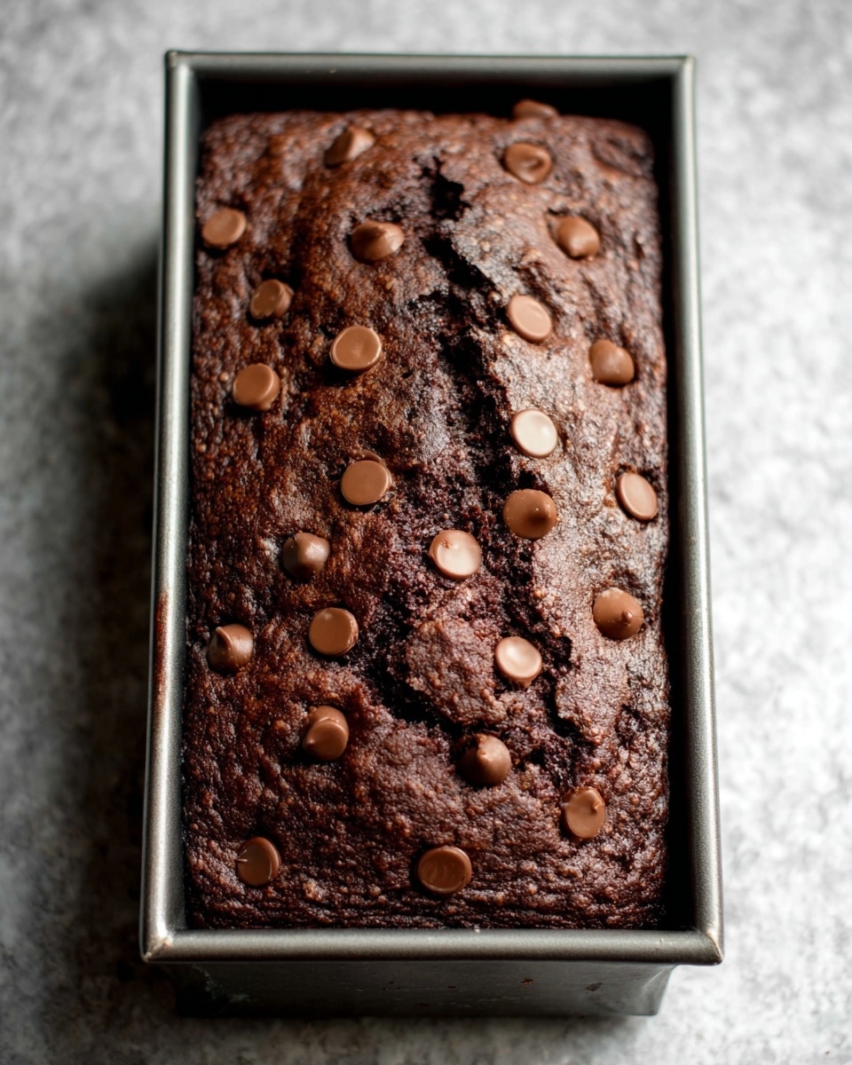 The image shows a thick chocolate loaf cake still in a metal baking pan. The cake has a dark brown, slightly cracked and moist-looking surface with a soft, dense texture. It is dotted evenly with light brown chocolate chips that are slightly melted into the cake's surface. The baking pan is sitting on a white marbled surface. The overall look is rich and dense, with a mix of smooth and rough textures visible on the cake's top photo taken with an iphone --ar 4:5 --v 7