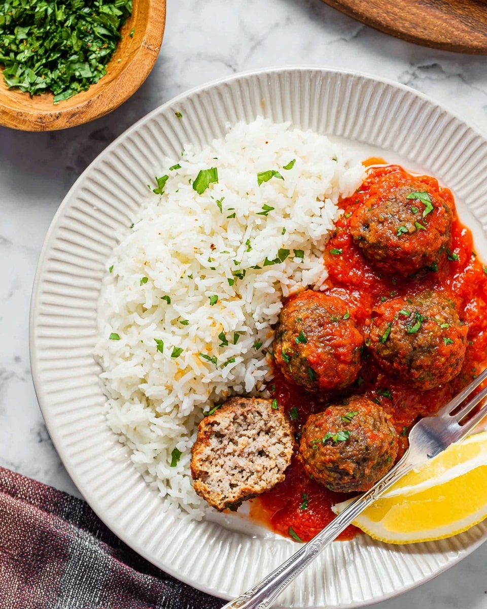 A white plate with ridged edges holds a serving of white rice on the left side, fluffy and separate grains visible. To the right, there are five round meatballs coated in a red tomato sauce, one meatball cut in half showing a moist, dense inside. The sauce beneath the meatballs is thick and chunky with bits of herbs sprinkled on top. A wedge of yellow lemon with a smooth peel rests at the bottom right corner of the plate. A metal fork is placed on the left side of the plate, its prongs resting on the tomato sauce near the rice. The setting includes a small wooden bowl filled with green chopped herbs in the upper left corner, all set on a white marbled surface. Photo taken with an iphone --ar 4:5 --v 7