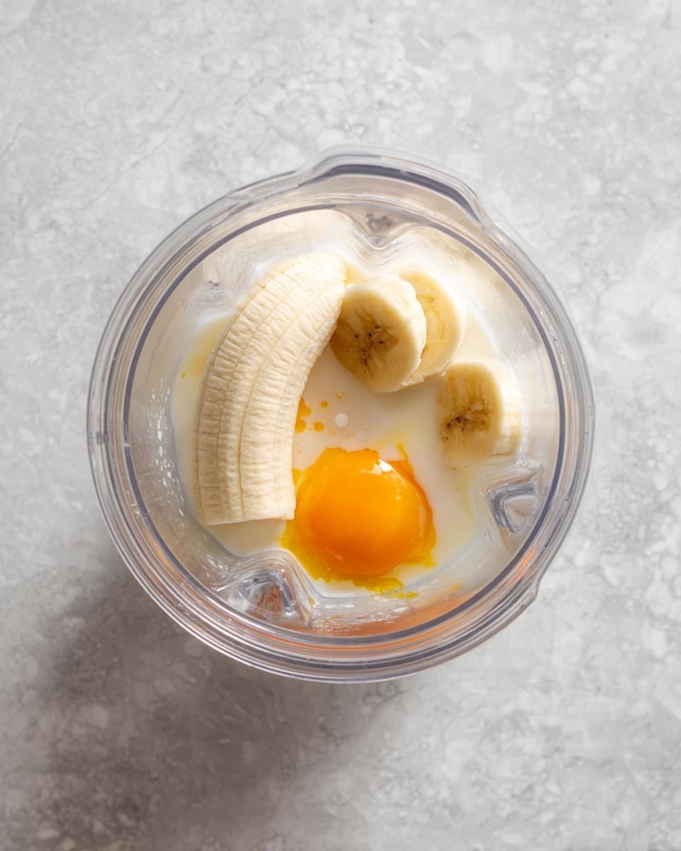 A clear blender cup is shown from above, containing three main ingredients: a half banana placed diagonally with its light yellow, smooth, and soft texture, an egg yolk with a bright orange color resting next to the banana, and a small amount of white milk pooling at the bottom. The blender cup is set against a white marbled textured surface. photo taken with an iphone --ar 4:5 --v 7