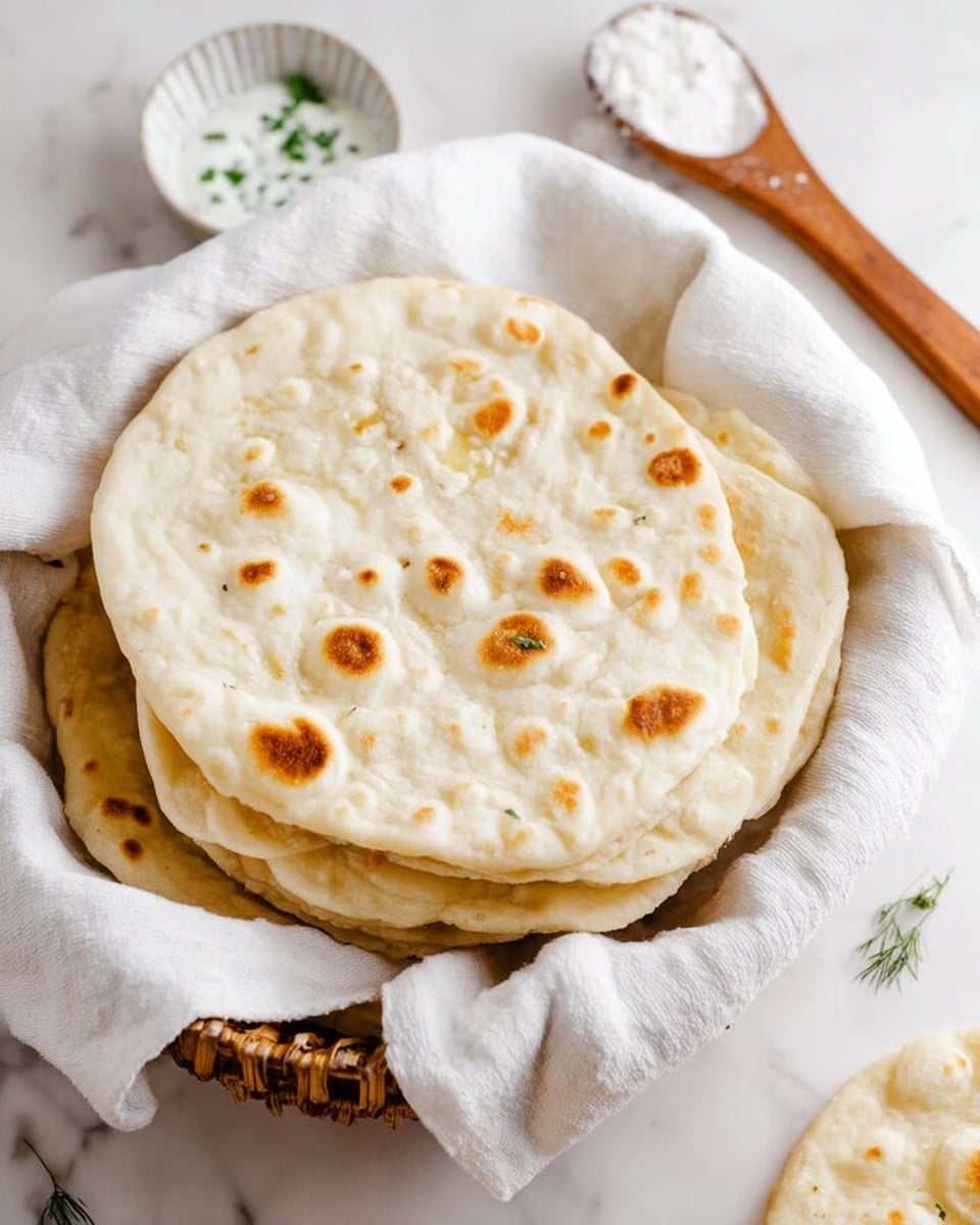 A basket lined with a soft white cloth holds a stack of four round flatbreads with light golden brown spots unevenly spread on their pale surface. Each flatbread is slightly puffy with a smooth texture and subtle bubbling. The basket sits on a white marbled surface next to a wooden spoon with some white flour on it and a small bowl of white sauce with herbs visible inside. Photo taken with an iphone --ar 4:5 --v 7