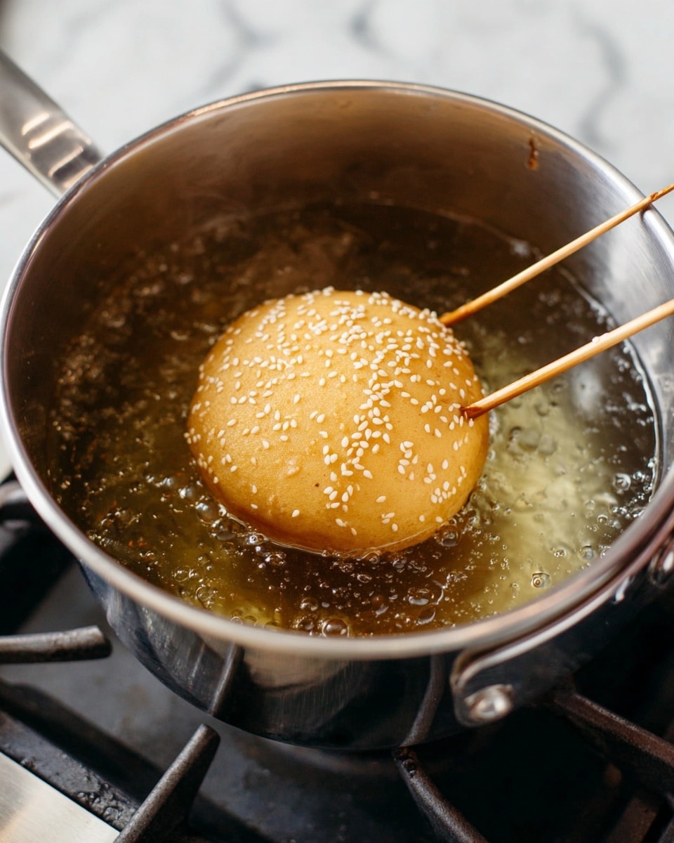 A golden-brown round dough ball with white sesame seeds on top is frying in a silver pot filled with hot oil. Two wooden sticks are holding the dough, one on the left and one on the top right, lifting it slightly above the oil. The silver pot sits on a stove burner with a white marbled surface visible in the background. The dough has a smooth texture with small bubbles on its surface, showing it is cooking. photo taken with an iphone --ar 4:5 --v 7