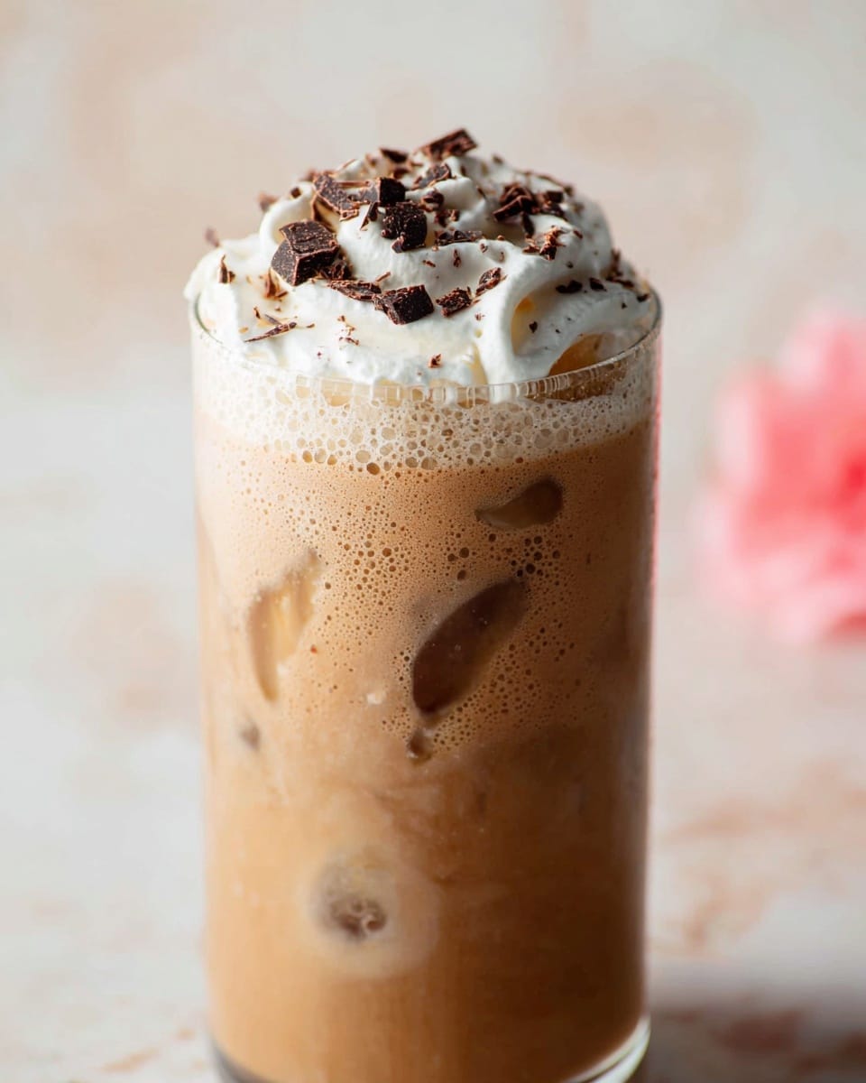 A tall clear glass filled with a creamy light brown iced coffee drink shows ice cubes inside. The drink has a frothy surface with small bubbles. On top, there is a thick swirl of white whipped cream sprinkled with dark brown chocolate shavings and chunks. The background is a soft white marbled texture with a pink blurry shape on the side. photo taken with an iphone --ar 4:5 --v 7