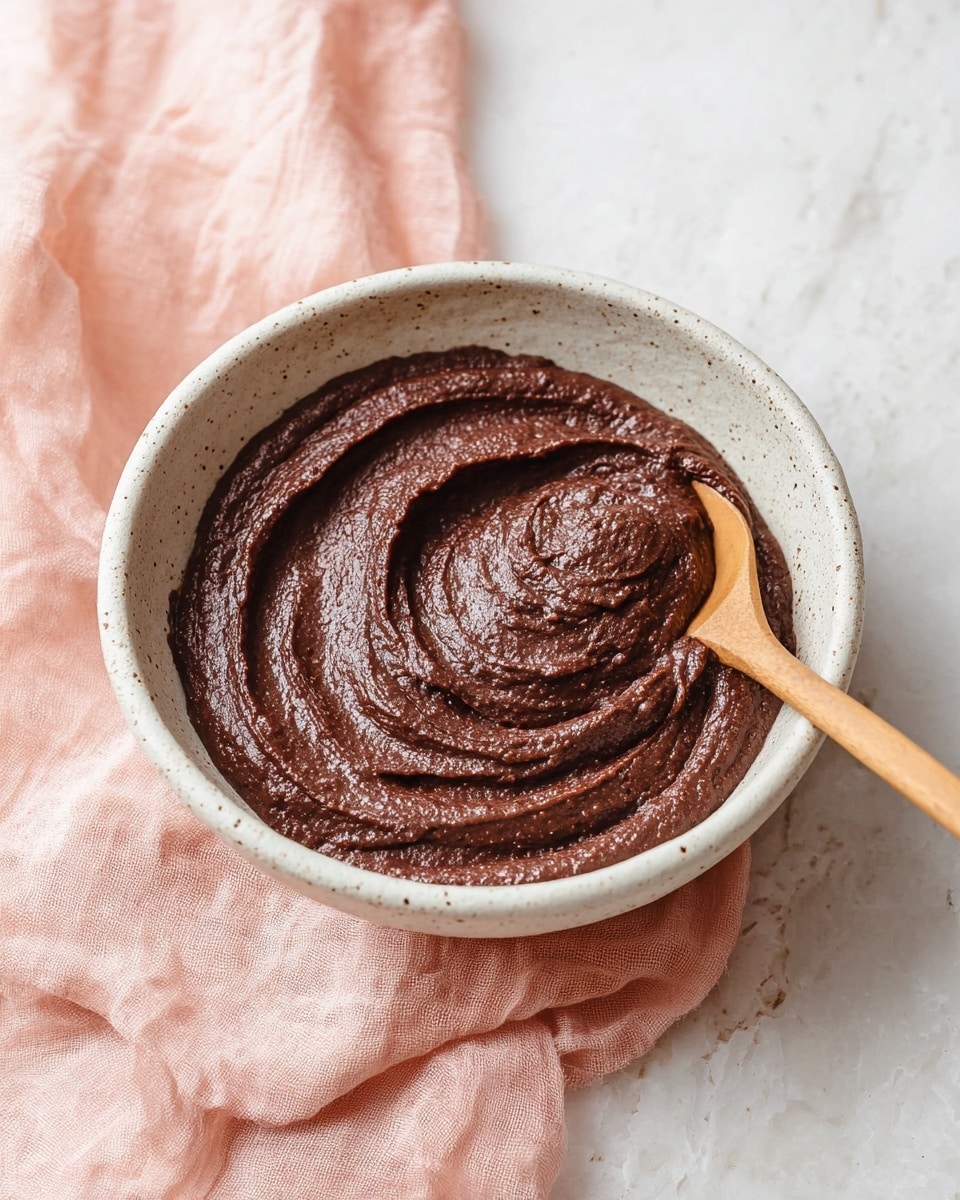 A white speckled bowl filled with smooth, thick, dark brown paste with a slightly grainy texture, swirled on top to show soft, creamy ridges. A thin wooden spoon rests on the right side, partially covered by the paste. The bowl sits on a soft, pale pink cloth placed on a white marbled surface. photo taken with an iphone --ar 4:5 --v 7