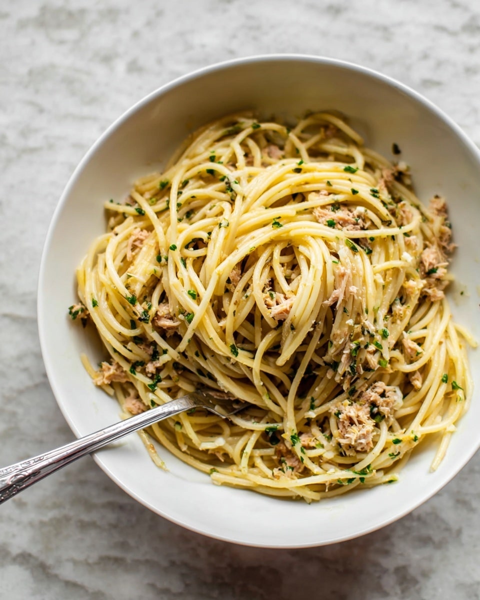 A white bowl filled with a generous serving of spaghetti noodles mixed with small pieces of tuna and sprinkled with chopped green herbs, creating a mix of pale yellow noodles and light brown tuna bits. A silver fork rests inside the bowl, twirling some of the spaghetti, with strands overlapping and intertwined, while the bowl sits on a white marbled surface. photo taken with an iphone --ar 4:5 --v 7