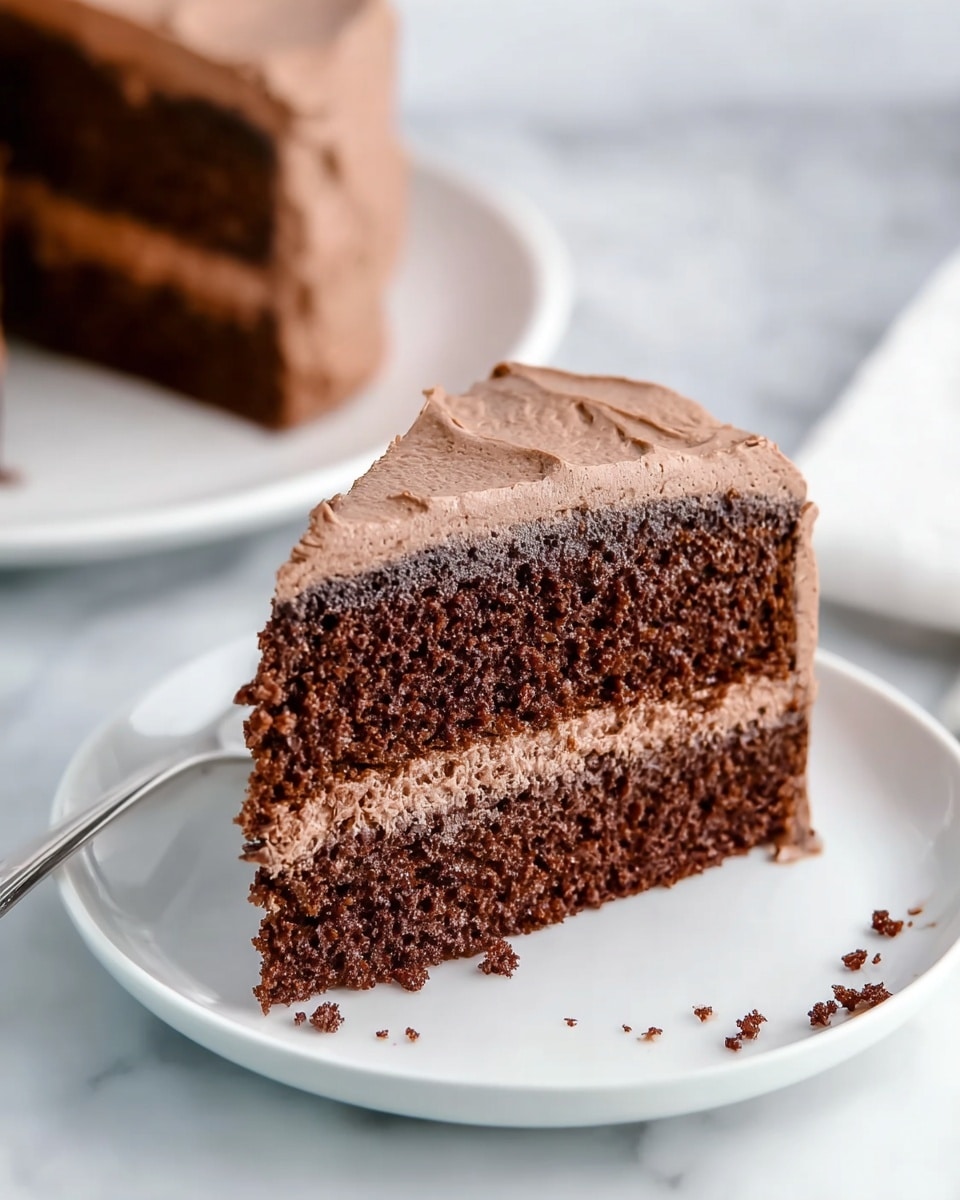 A slice of chocolate cake with two thick layers of dark brown, moist cake separated by a smooth, light brown chocolate frosting layer in the middle. The outside of the slice is covered with the same light brown frosting that looks creamy and soft. It sits on a clean white plate on a white marbled surface, and there are some small crumbs near the bottom right of the slice. Another slice is blurred in the background on a white plate. Photo taken with an iphone --ar 4:5 --v 7