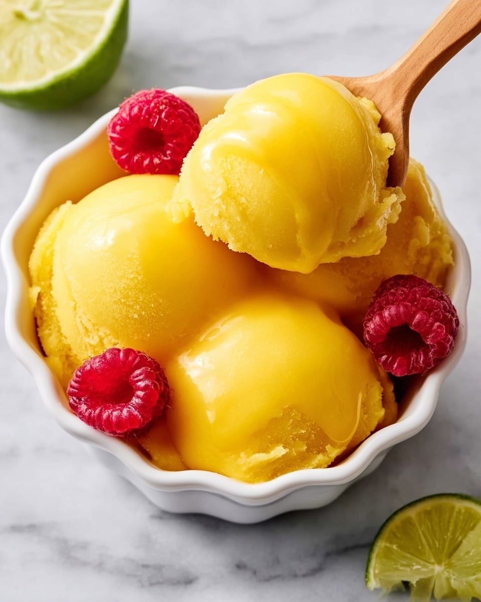A white scalloped bowl is filled with three bright yellow scoops of smooth mango sorbet, each scoop rounded and glossy. Two fresh red raspberries sit beside the sorbet, adding a pop of vibrant color. A wooden spoon is lifting a scoop of the sorbet, showing its creamy texture. Near the bowl, there is a green lime wedge adding a fresh touch. The whole scene is set on a white marbled surface. photo taken with an iphone --ar 4:5 --v 7
