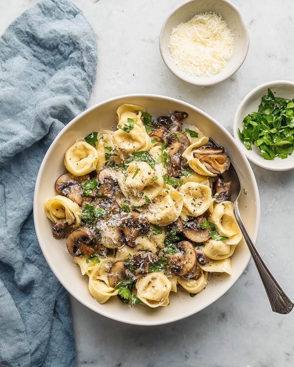 A white bowl filled with about three layers of tortellini pasta having a pale yellow color and soft texture. Mixed in are sliced sautéed brown mushrooms with a slightly shiny surface. Green parsley leaves are scattered within and under the pasta, adding a fresh green touch. The dish is lightly sprinkled with finely grated white cheese and ground black pepper on top. A silver spoon rests inside the bowl on the right side. The bowl sits on a white marbled surface with a small white bowl of grated cheese above and a small white bowl of green herbs to the right. A soft blue cloth lies to the left of the bowl. The lighting is natural and bright. Photo taken with an iphone --ar 4:5 --v 7