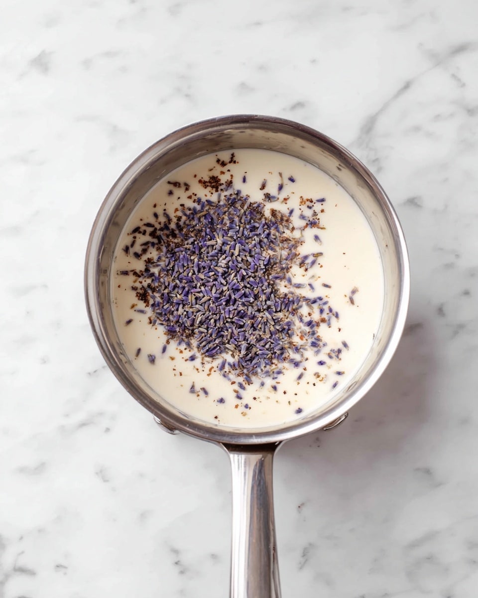A silver metal saucepan sits on a white marbled surface, filled halfway with a smooth, light cream-colored liquid. On top of the liquid, there is a scattering of small, dried purple and brown lavender flowers mostly concentrated in the center, creating a contrast of soft colors against the creamy base. The saucepan handle points to the bottom right corner of the image. photo taken with an iphone --ar 4:5 --v 7