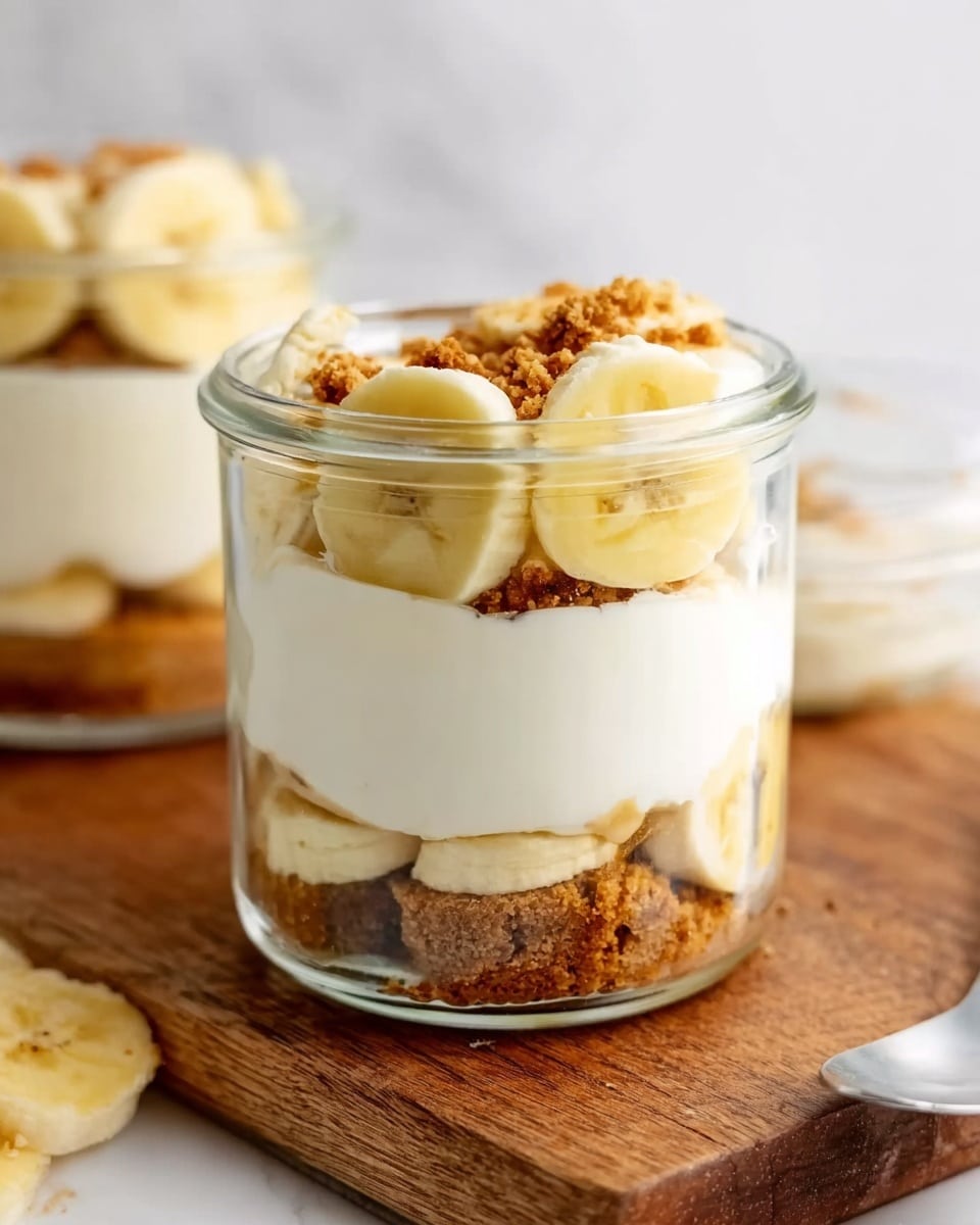 The image shows a clear glass jar with three main layers. The bottom layer has light brown pieces that look crunchy and crumbly. The middle layer is thick and white, smooth and creamy in texture. The top layer holds slices of yellow banana arranged around the edge and more white cream filling the center, with some brown crunchy bits sprinkled on top. The jar sits on a wooden surface with a white marbled background. Another similar jar is seen blurred in the background. Photo taken with an iphone --ar 4:5 --v 7