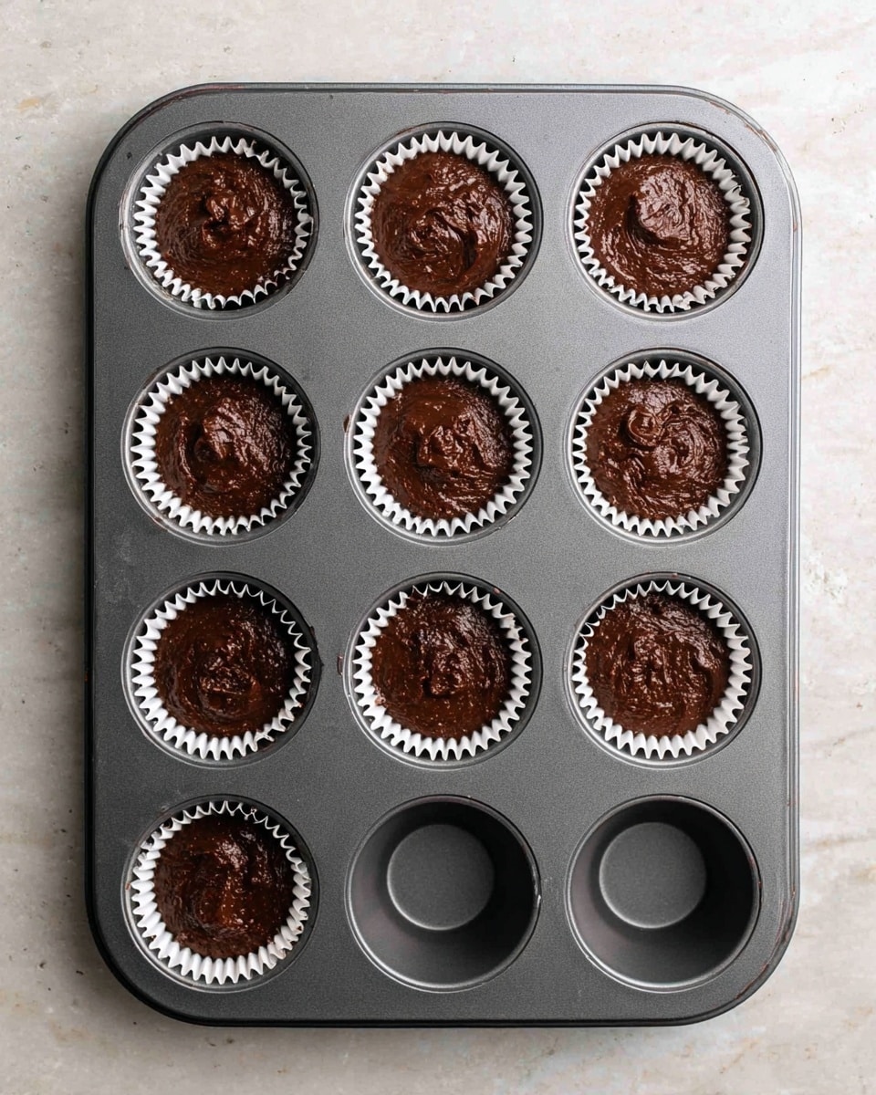 A dark gray metal muffin tray with twelve cup slots sits on a white marbled surface, five of the cups lined with white paper liners filled with dark brown, thick, shiny chocolate batter. The batter has a slightly rough texture with small uneven peaks and some smooth areas, filling less than half of each liner. The empty slots show a clean, matte gray metal interior. The tray is seen from above, centered in the image. photo taken with an iphone --ar 4:5 --v 7