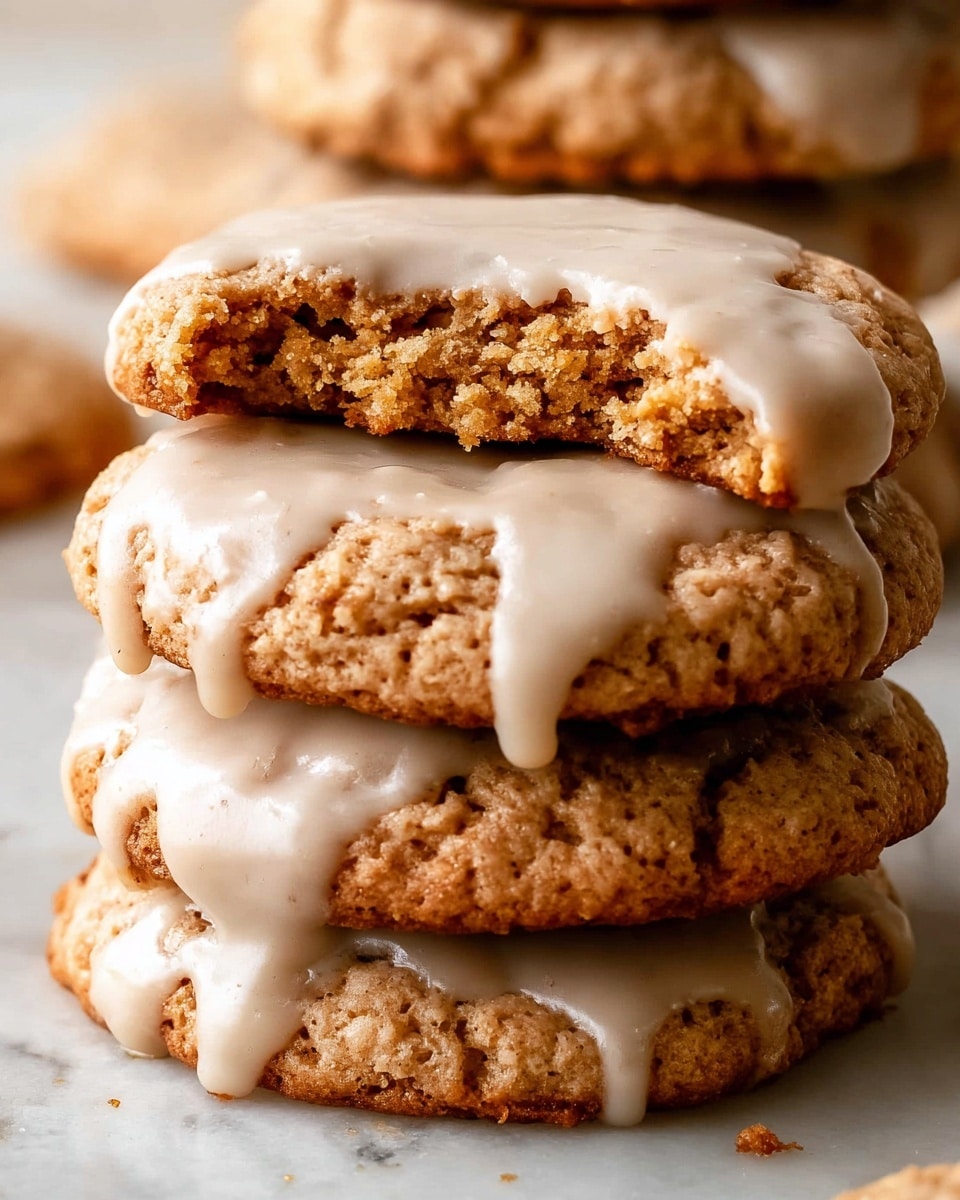 A close-up view of a stack of oatmeal cookies with light brown icing dripping down the edges. The bottom stack has three cookies, each with a rough, slightly crumbly texture, and the top cookie shows a bite taken out, revealing a dense, chewy inside. The icing on each cookie is smooth and glossy, pooling slightly on the sides. Another stack of similarly iced cookies stands blurred in the background on a white marbled surface. photo taken with an iphone --ar 4:5 --v 7