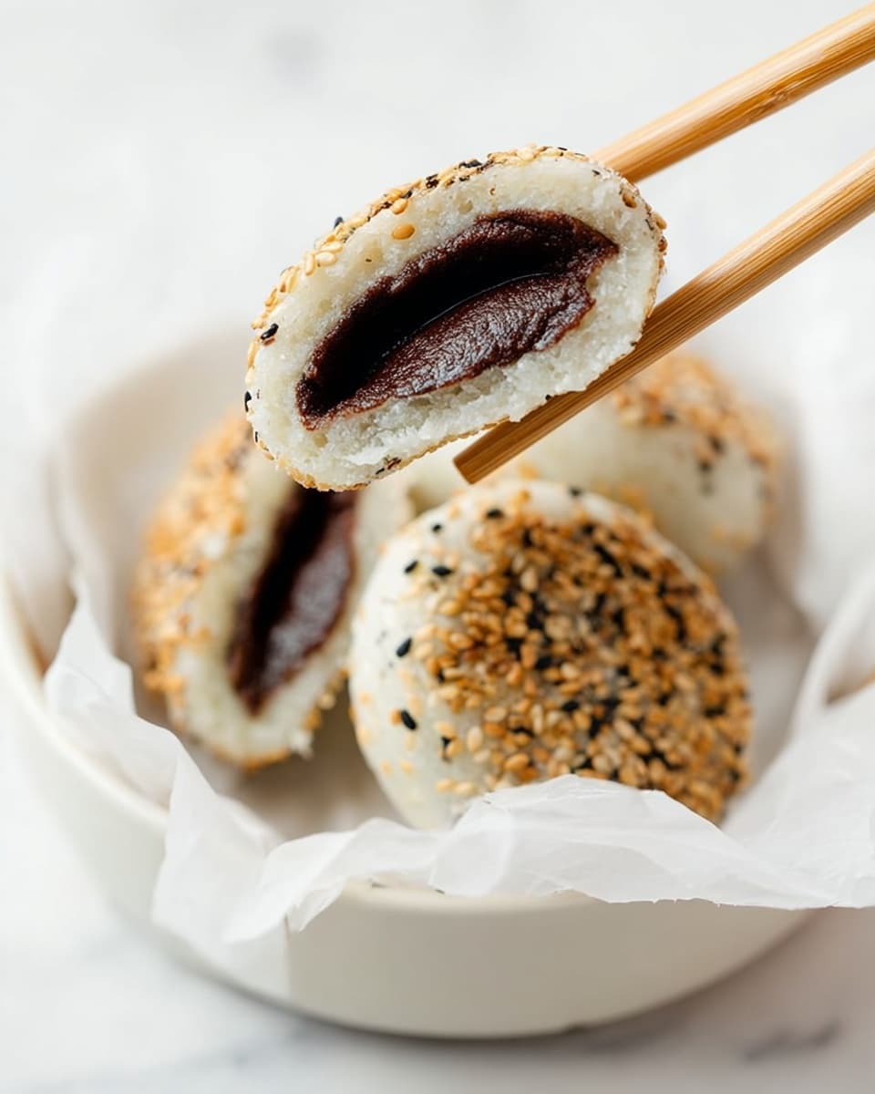 The image shows a close-up of a white bowl lined with white parchment paper, holding three round, sesame-coated snacks. One snack is held by wooden chopsticks, revealing its inside layers: a soft white outer layer dusted with toasted sesame seeds and a dark, smooth red bean filling in the center. Two more snacks sit in the bowl, showing their golden-brown toasted sesame seed tops against the white parchment paper and bowl, all set on a white marbled surface. Photo taken with an iphone --ar 4:5 --v 7