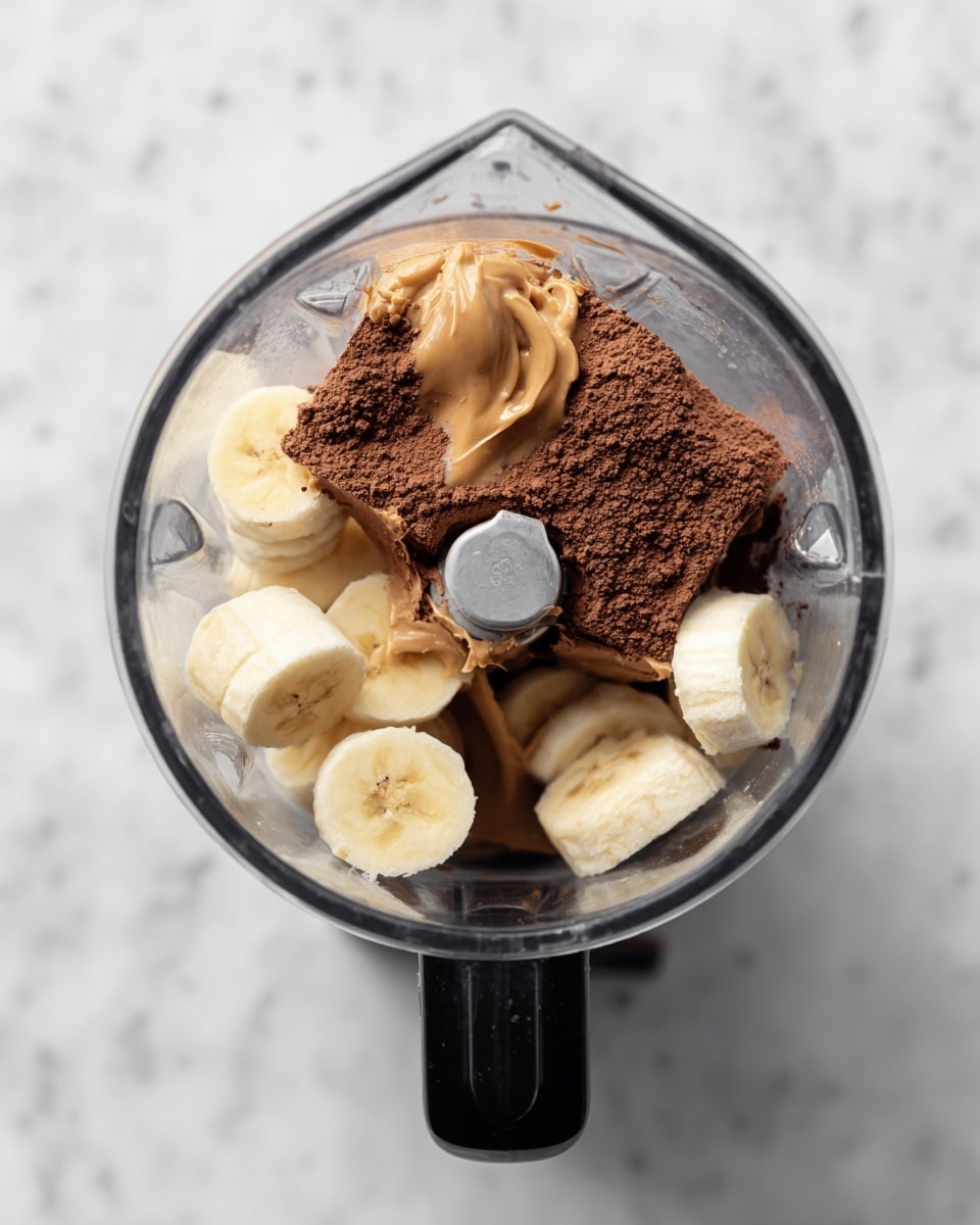 The image shows a blender jar viewed from above on a white marbled surface. Inside the blender, there are three main layers of ingredients: at the bottom, there are round slices of banana with a pale yellow color and soft texture; on top of the banana slices is a dollop of creamy light brown peanut butter; covering one side of the peanut butter is a heap of fine, dark brown cocoa powder. The blender blade is in the center, with some cocoa powder sticking to one side of it. The jar is clear and the handle is black. photo taken with an iphone --ar 4:5 --v 7