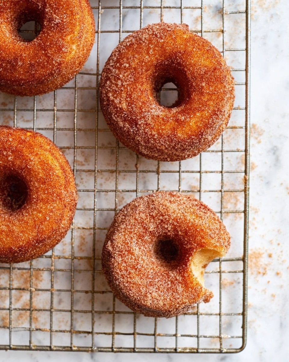 Four cinnamon sugar-coated donuts are placed on a metal cooling rack over a white marbled surface. Each donut has a golden-brown color with a rough sugar texture on the outside. One donut near the center has a bite taken out of it, showing a soft, light inside. The sugar coating is unevenly spread, giving a natural homemade look, and some sugar has fallen onto the cooling rack and surface below. Photo taken with an iphone --ar 4:5 --v 7