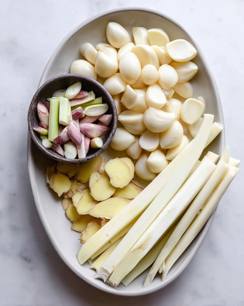 An oval white bowl sits on a white marbled surface and holds several layers of ingredients. The top right side is filled with many peeled garlic cloves, smooth and off-white. To the bottom right, white elongated lemongrass stalks are laid side by side. The bottom left has peeled ginger root pieces, light yellow with rough edges. On the top left inside the bowl, a small round dark bowl holds thin slices of pale green and light purple lemongrass pieces. The colors are soft with natural textures, showing clean and fresh ingredients. Photo taken with an iphone --ar 4:5 --v 7