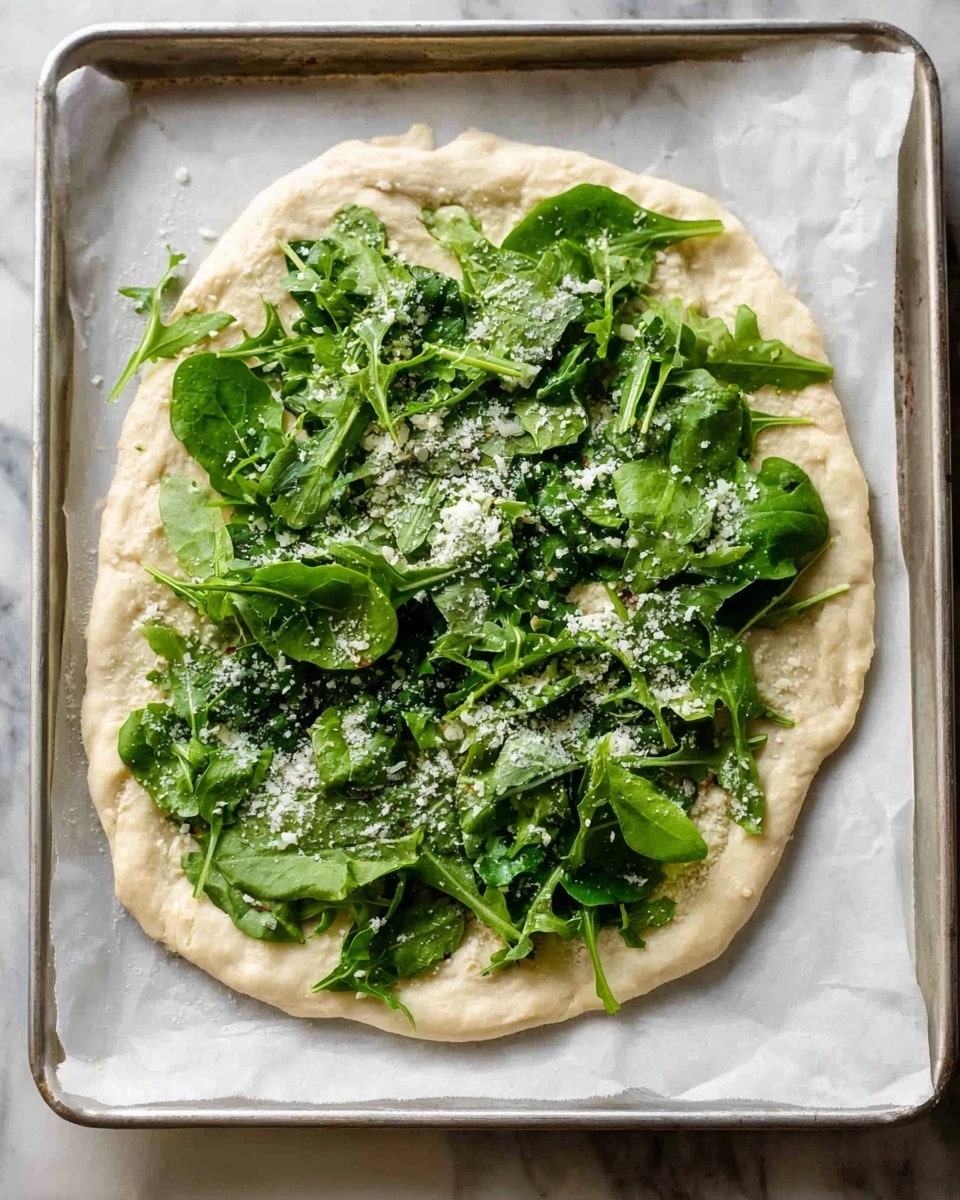 The image shows an uncooked pizza on a baking tray lined with white parchment paper. The crust is thick and irregularly shaped with a pale beige dough base. On top of the dough, there is a layer of fresh, dark green leafy vegetables, including spinach and arugula, scattered unevenly but covering most of the surface. Over the greens, there is a sprinkling of white grated cheese, giving a slightly powdery texture. The baking tray is silver metal and sits on a white marbled surface. Photo taken with an iphone --ar 4:5 --v 7