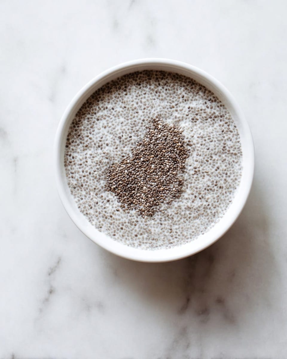 A white round bowl filled with a single layer of grayish-white chia seed pudding with tiny dark seeds evenly spread throughout the surface. The texture looks soft and slightly thick, with a small uneven clump in the center. The bowl is placed on a white marbled surface. photo taken with an iphone --ar 4:5 --v 7