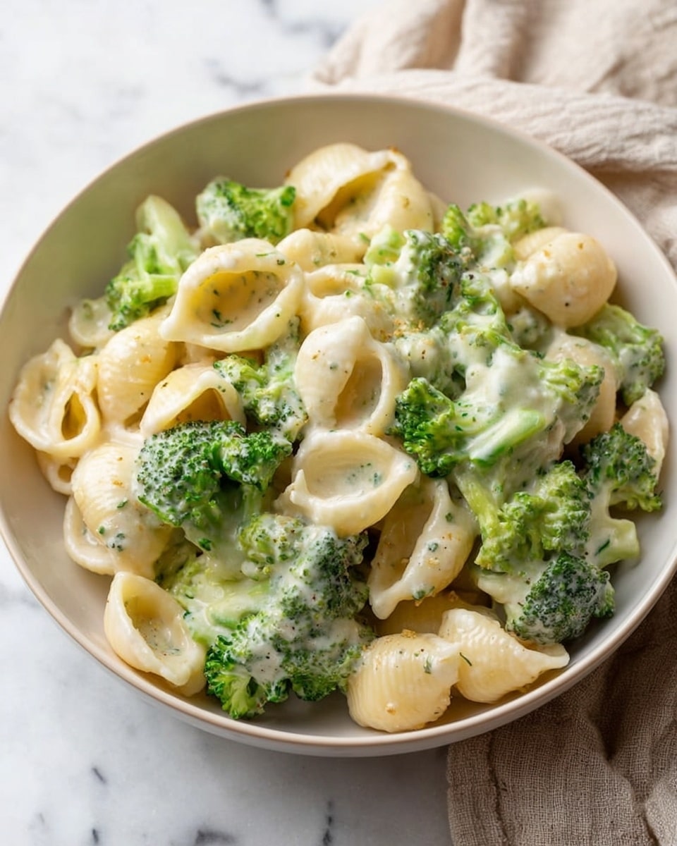 A white bowl filled with small curved pasta shells mixed with bright green broccoli florets. The pasta and broccoli are covered in a thick creamy white sauce that has a smooth texture and slight specks of herbs. The shells are soft with a slightly glossy finish, and the broccoli pieces look tender yet firm. The bowl sits on a white marbled surface with a light beige cloth partially visible nearby. photo taken with an iphone --ar 4:5 --v 7