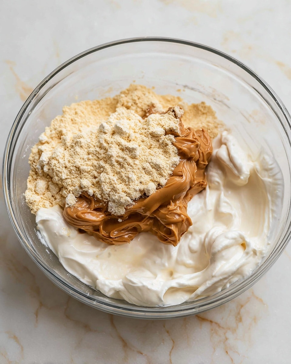 A clear glass mixing bowl holds three main layers of ingredients. The bottom layer is a smooth, white creamy base with soft peaks and fluffy texture. On top, there is a thick, glossy brown peanut butter layer, slightly swirled and soft looking. The top layer is a loose pile of pale yellowish powder, scattered unevenly over the peanut butter. The bowl sits on a white marbled surface that has faint grey veins. The photo taken with an iphone --ar 4:5 --v 7