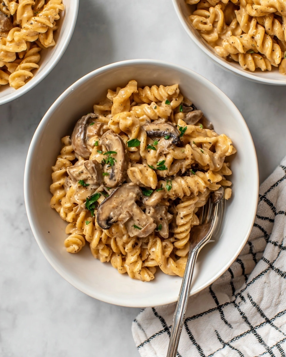 A white bowl sits on a white marbled surface filled with creamy pasta made of tan twisted rotini noodles and sliced light brown mushrooms mixed evenly throughout. The pasta is creamy and covered with a smooth light brown sauce, sprinkled with small bits of fresh green herbs and black pepper. A shiny silver fork rests inside the bowl on the right side, touching the pasta. Part of two other white bowls with the same pasta are partially visible at the top edges of the image. A white cloth with black grid lines is placed near the bottom right corner. photo taken with an iphone --ar 4:5 --v 7