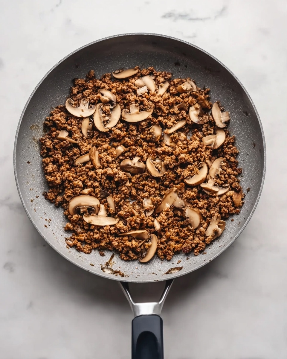 A grey frying pan with a black handle is filled with cooked ground meat mixed with thin, light brown mushroom slices spread evenly throughout. The mixture is crumbly with some small uneven clumps, showing a mix of dark brown and lighter brown colors. The pan is placed on a white marbled surface. photo taken with an iphone --ar 4:5 --v 7