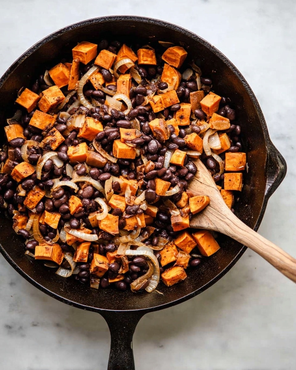 A black cast iron pan is filled with a mix of small cubed orange sweet potatoes and dark brown black beans, with thin, light beige slices of cooked onions scattered throughout. The sweet potatoes and black beans are mixed evenly, showing a slightly crispy texture on some cubes. A wooden spatula rests inside the pan, partially buried under the mixed ingredients. The pan sits on a white marbled surface. photo taken with an iphone --ar 4:5 --v 7