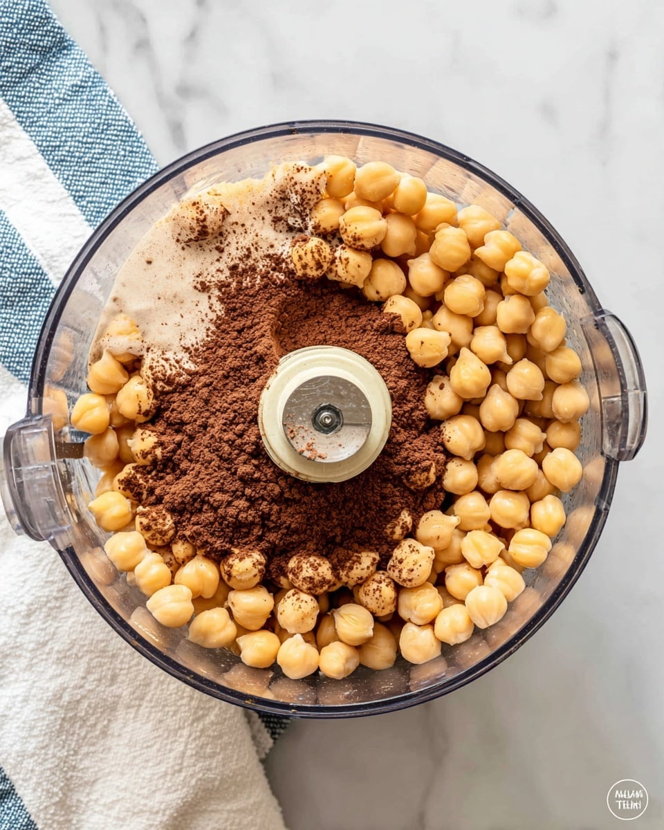 The image shows a clear food processor bowl filled with three main layers. The bottom layer is made of light tan chickpeas evenly spread around the bowl. Above the chickpeas, there is a light brown liquid layer visible through the chickpeas. On top of the liquid, there is a dark brown powder of cocoa or cacao placed mostly in the center. The bowl is placed on a white marbled surface with a white towel featuring blue stripes partly visible on the left side. The food processor blade and mechanism are seen in the middle center of the bowl. Photo taken with an iphone --ar 4:5 --v 7