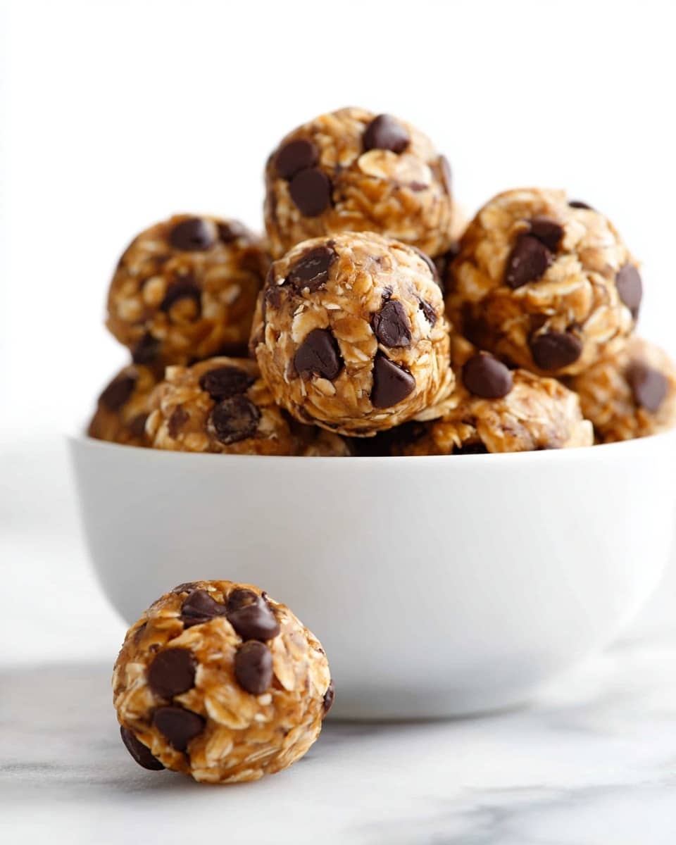 A white bowl filled with small round energy bites that have a light brown oatmeal texture mixed with dark brown chocolate chips spread throughout each bite. The energy bites appear soft and chunky, stacked closely inside the bowl, with two bites placed outside on a white marbled surface in the foreground. The focus is sharp on the bites, showing detailed oat flakes and chocolate chips with bright lighting against a clean white background. photo taken with an iphone --ar 4:5 --v 7