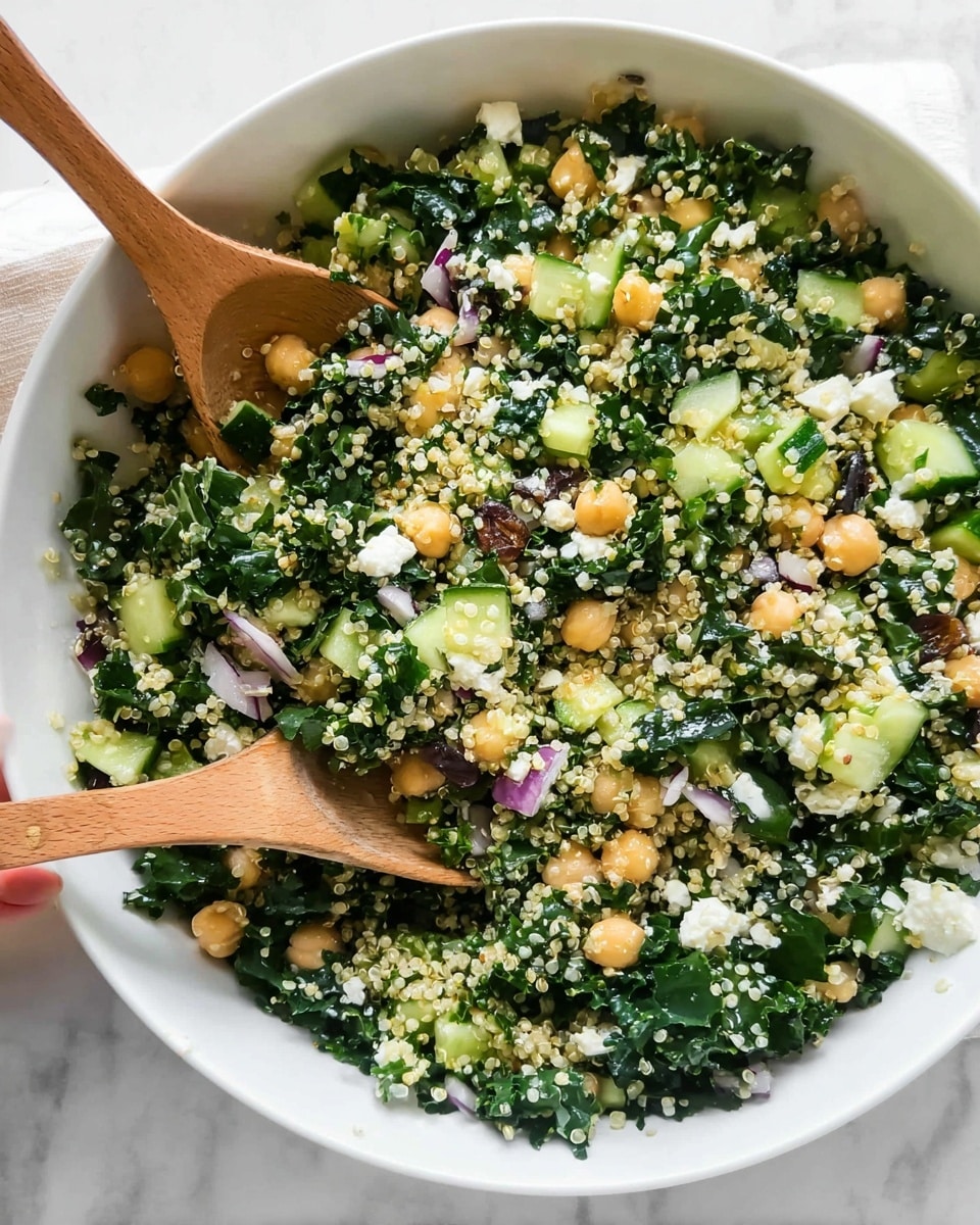 A white bowl filled with a fresh mixed salad showing layers of small green quinoa grains scattered evenly throughout large pieces of dark green kale leaves, light green cucumber chunks, pale beige chickpeas, small white crumbled cheese, tiny golden raisins, and small diced pieces of purple onion. Two wooden spoons rest inside the bowl, one being held by a woman's hand from the left side. The bowl sits on a white marbled surface, highlighting the colorful and textured mixture of the salad photo taken with an iphone --ar 4:5 --v 7