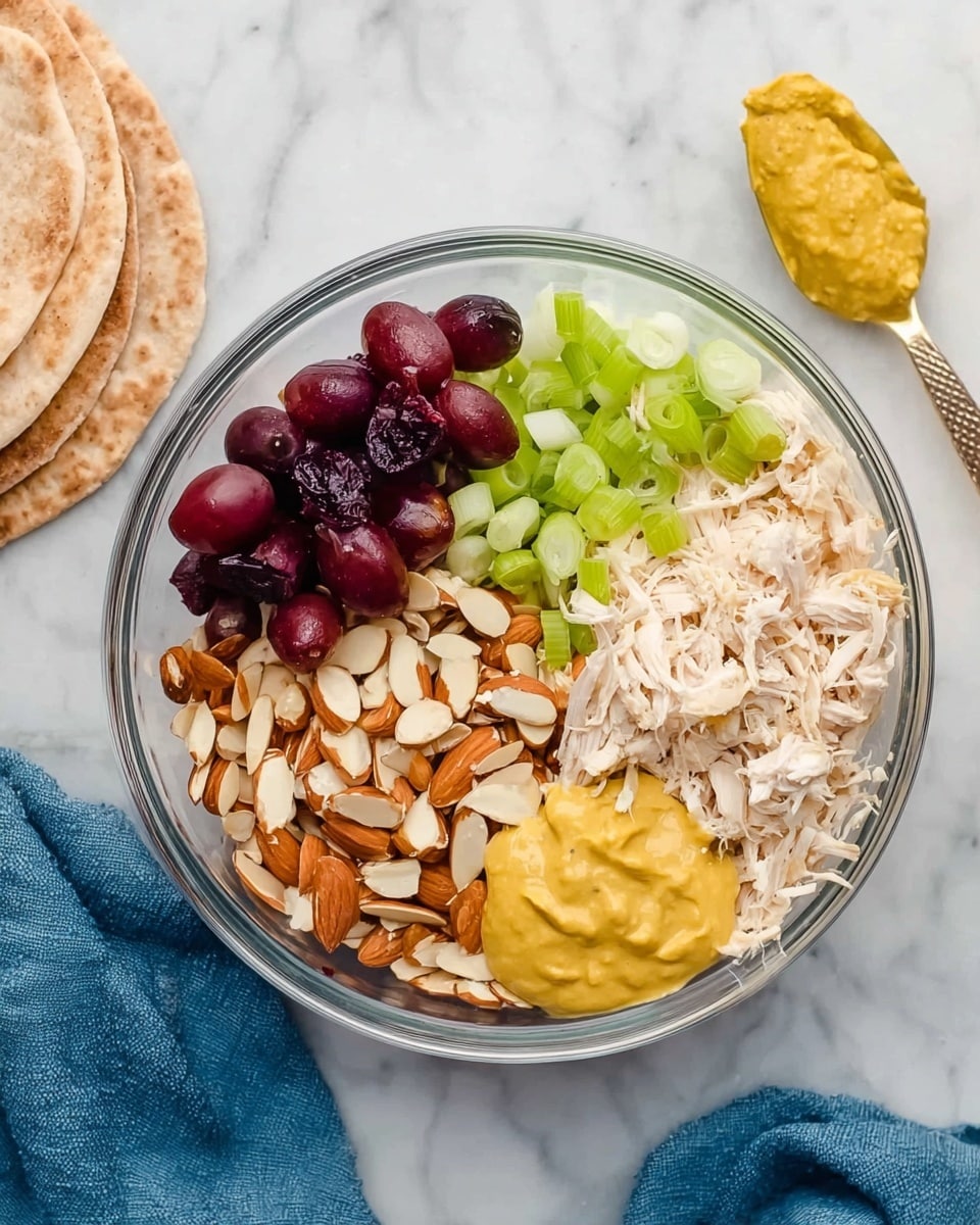 A clear glass bowl on a white marbled surface holds a layered mix of ingredients. On the top left, there are dark red grape halves, below them is a pile of small, thinly sliced green onions. Next to the onions, on the bottom center, is a heap of sliced almonds with their skins on. On the top right, shredded white chicken meat fills the space, while on the far right side, a smooth yellow mustard-like sauce sits in a spooned shape. Next to the bowl on the left side, there are three pieces of flatbread stacked slightly overlapping. A blue cloth is partially visible on the right side of the image. Photo taken with an iphone --ar 4:5 --v 7