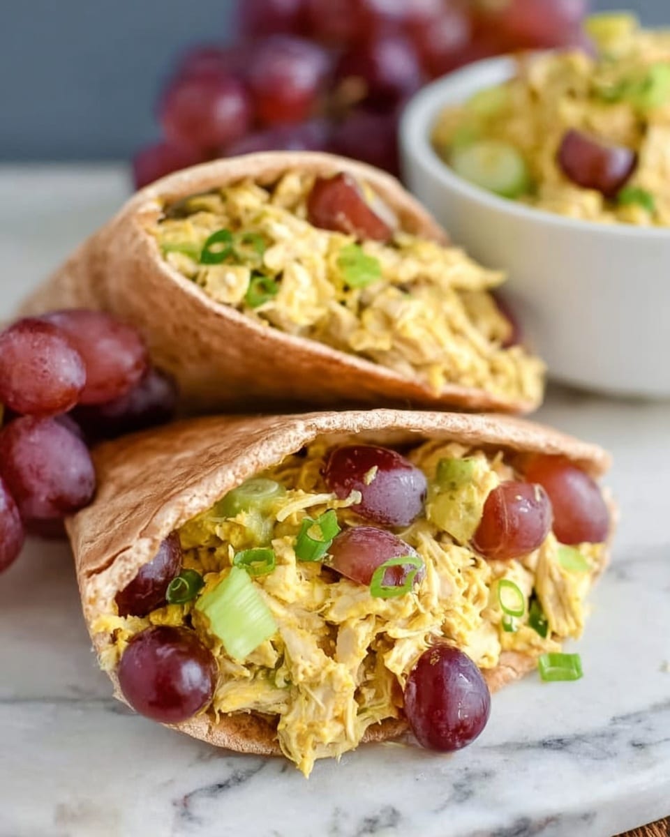 The image shows two pita pocket halves filled with shredded yellow chicken mixed with green onions and sliced red grapes, giving a colorful and fresh look. The pita bread is light brown and slightly toasted, holding the filling firmly. In the background, there is a white bowl with more of the same chicken mixture, and a cluster of red grapes sits on the white marbled surface beside the pita pockets. The overall look is bright and fresh, with the soft textures of the chicken and grapes contrasting with the slightly crispy pita. Photo taken with an iphone --ar 4:5 --v 7