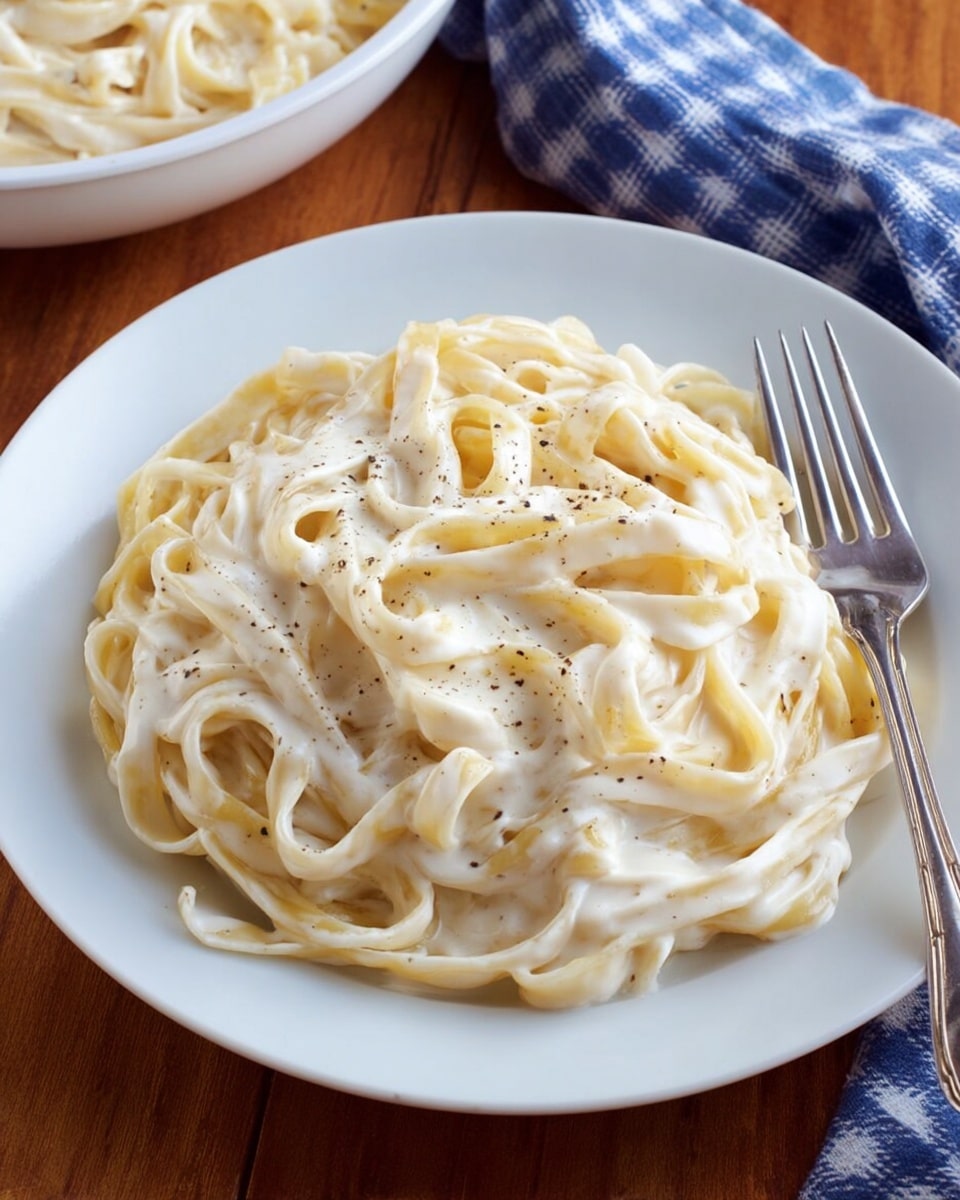 A white round plate with a nest of creamy fettuccine pasta coated in a smooth, thick white sauce, lightly speckled with black pepper. The pasta strands are loosely twisted with a soft texture, filling the center of the plate. To the right side of the plate, there is a silver fork resting on the edge. In the top left corner of the image, a white bowl is partially visible, showing more of the saucy pasta inside. The background features a wooden table surface with a blue and white checkered cloth beside the plate. photo taken with an iphone --ar 4:5 --v 7